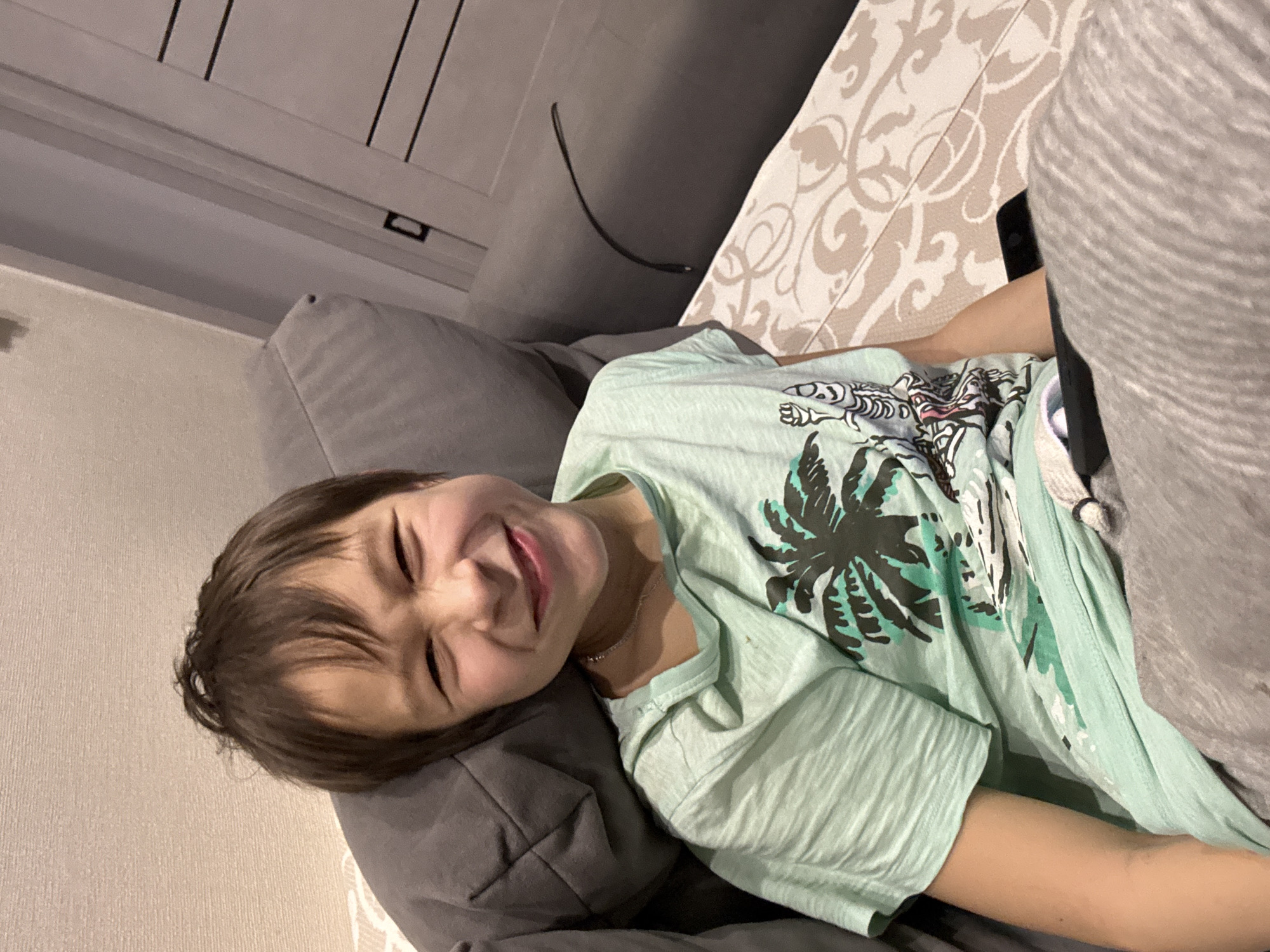 A boy is lying on a gray sofa. He is wearing a light green t-shirt with a palm tree design. He is smiling and squinting his eyes. Part of a table with a patterned surface is visible next to the sofa. A black cable is hanging down from above. The location is assumed to be in a domestic setting, as per the image's origin in Chaikovsky, Russia.
