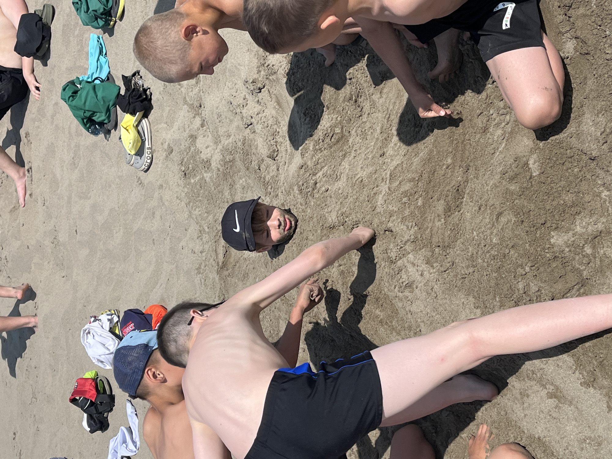 The image depicts a group of young boys playing on a sandy beach. One boy is partially buried in the sand, with only his head and shoulders visible. He is wearing a black baseball cap with a white Nike logo. Two other boys are kneeling and interacting with the sand, seemingly burying their friend or playing in the sand. Another boy is visible in the background, also on the beach. Scattered clothing and footwear are present on the sand. The environment is a beach with light brown sand and visible shadows suggesting a sunny day. The scene is consistent with a leisure activity at a beach, possibly near the city of Krasnoturansk, Russia, given the context.