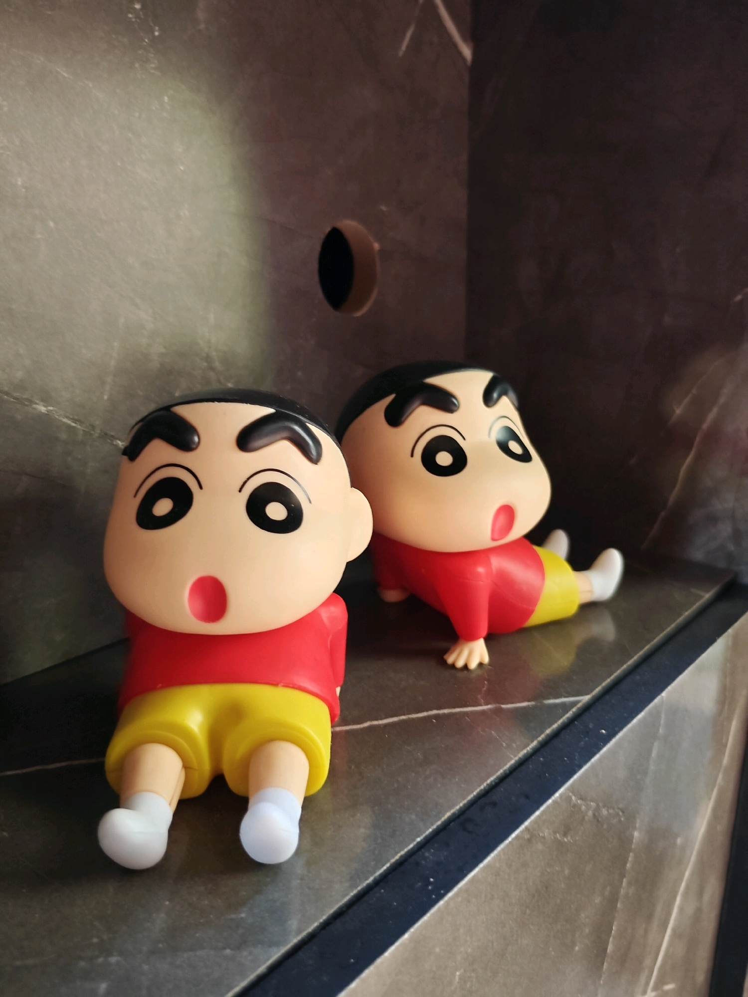 Two Shin-chan dolls are positioned on a dark, textured surface. The doll on the left is sitting upright, while the doll on the right is in a crawling position. Both dolls are wearing red shirts and yellow shorts. The background features a dark, marbled wall with a circular cutout. The scene appears to be indoors, possibly on a shelf or countertop. There is no discernible connection to Phra Nakhon Si Ayutthaya City Municipality, Thailand, as the image lacks any identifying landmarks or context specific to that location.