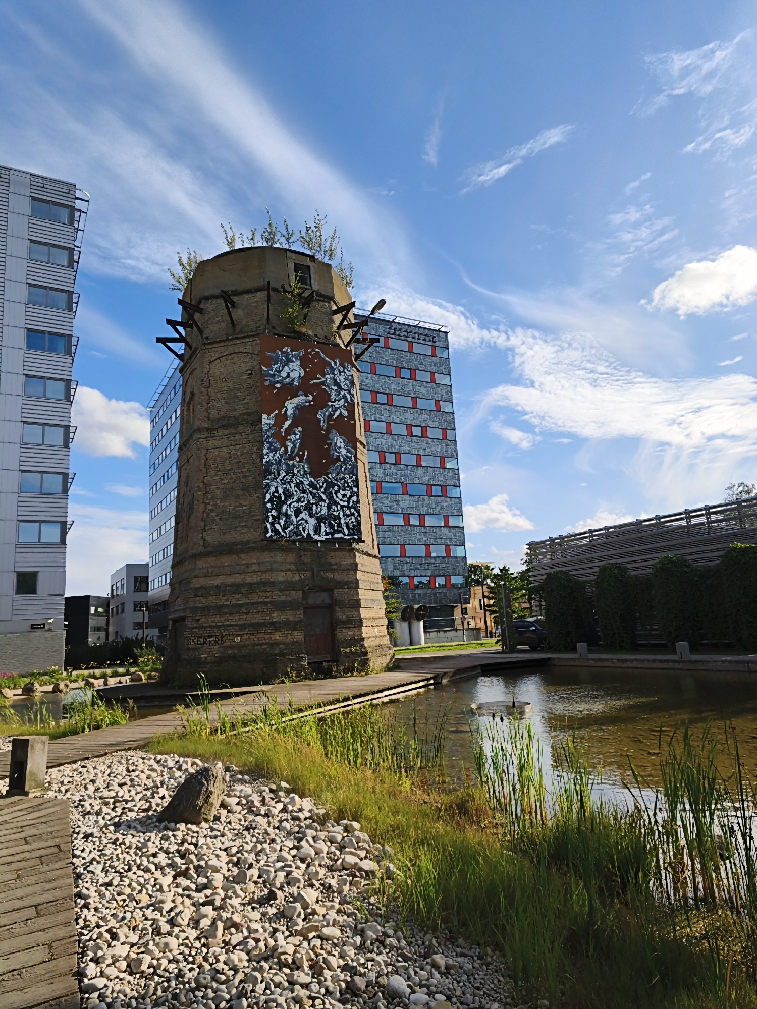 The image shows a brick tower, possibly an old industrial chimney, adorned with a large mural depicting a chaotic scene of figures in black and white against a reddish-brown background. The tower is situated in an urban environment with modern buildings in the background, one of which is a tall, glass-fronted structure with red window frames. A body of water, likely a pond or canal, is in the foreground, with reeds and grassy banks. Wooden walkways and a gravel path with a prominent rock are also visible. The sky is blue with wispy white clouds. The overall scene suggests a blend of industrial heritage and modern urban development. The location is the Telliskivi Creative City in Tallinn, Estonia. There are no people visible in the image.