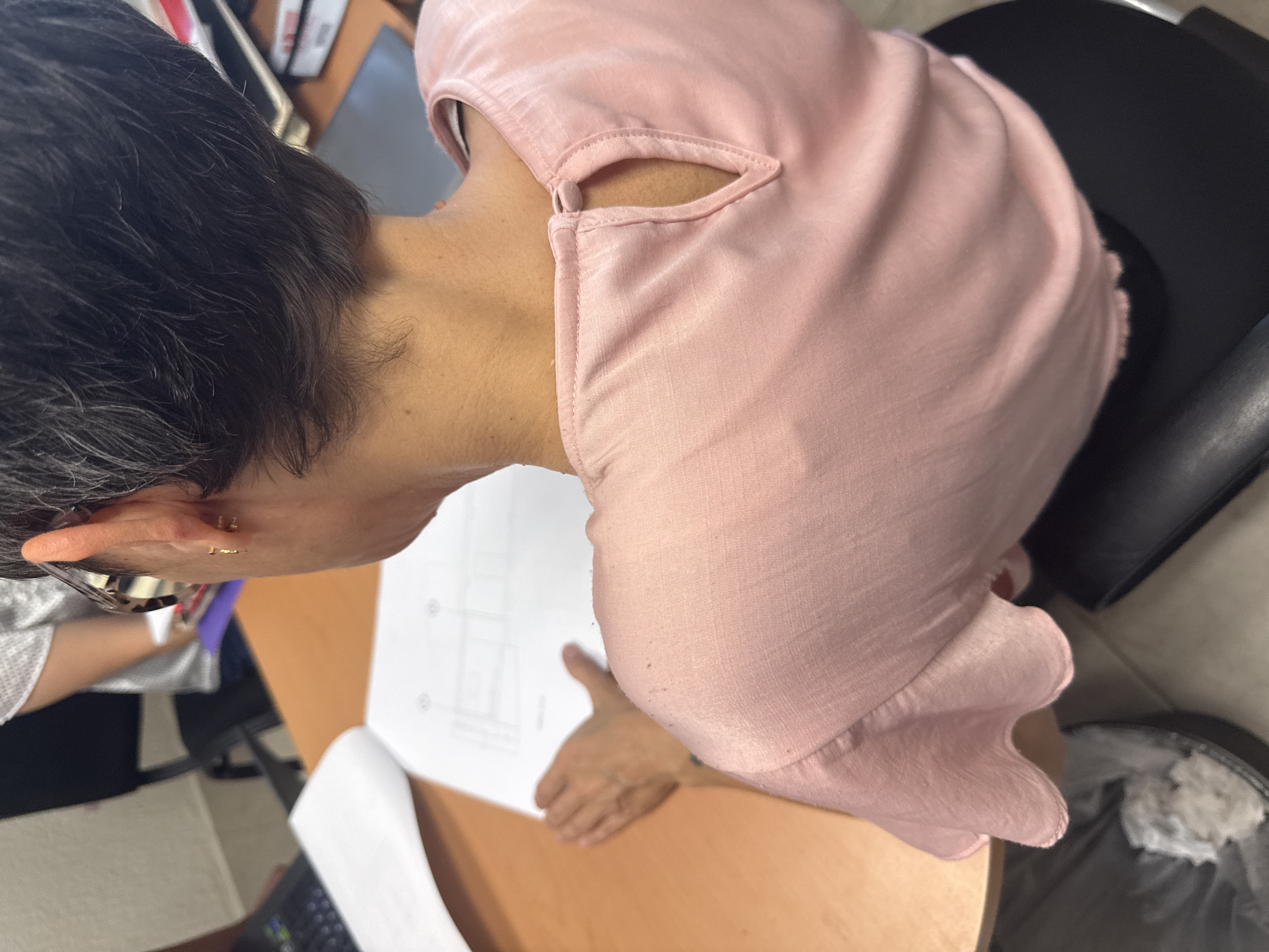 A person with short, dark hair and glasses is looking down at a piece of paper with technical drawings on a light brown desk. The person is wearing a light pink, loose-fitting top with a cutout at the neckline. The desk is cluttered with other papers, books, and office supplies. A black office chair is visible to the right of the person. The location is San Andrés Cholula, Mexico.