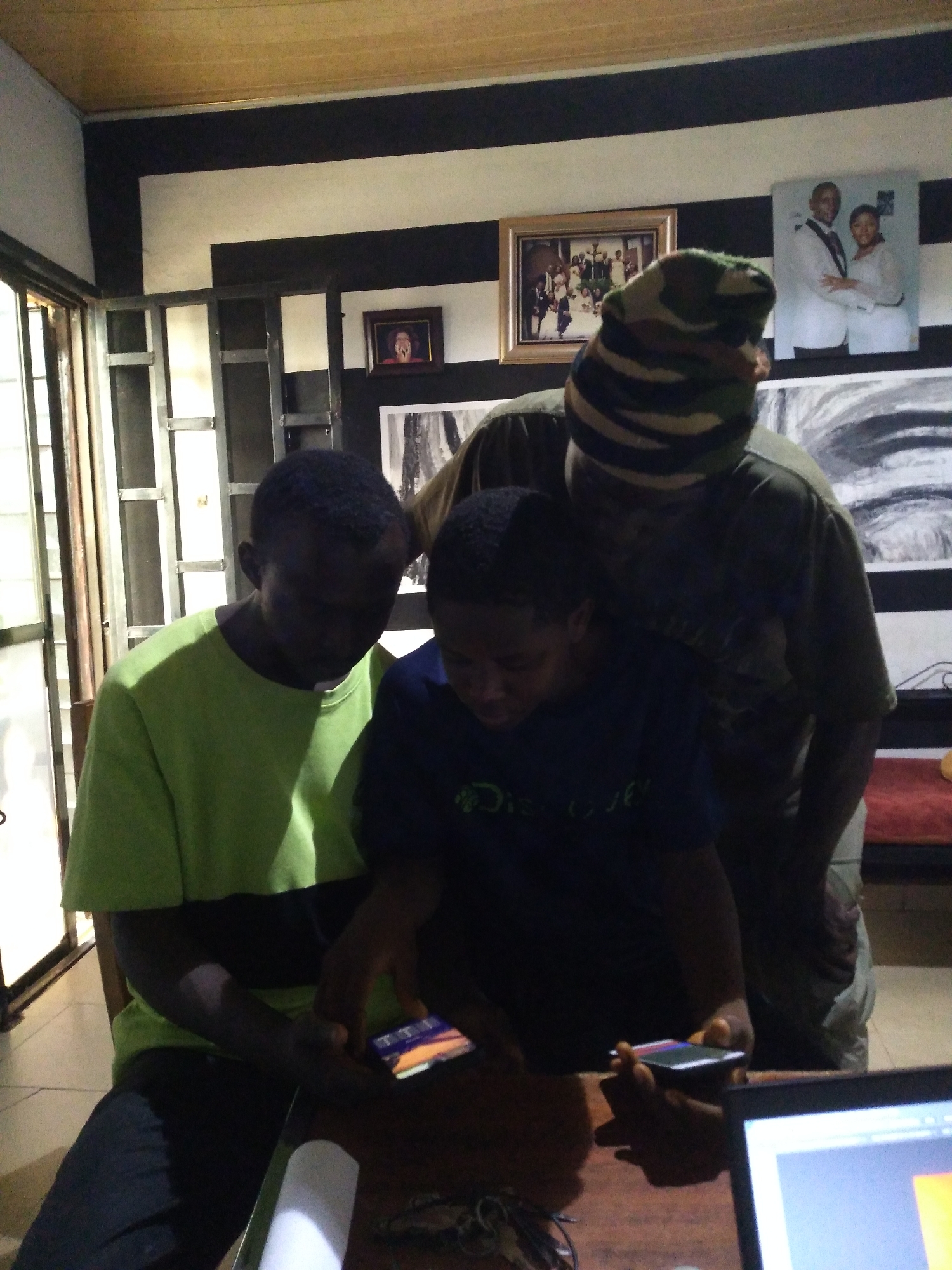 Three individuals are gathered around a table, two of whom are looking at a smartphone held by the person in the foreground. The person in the foreground is wearing a bright green t-shirt and dark pants. The person behind them is wearing a dark t-shirt and a camouflage beanie. The third individual, partially obscured, is leaning over the table. The scene appears to be indoors, with a distinctive black and white striped wall in the background and a framed picture on the wall. The presence of a laptop screen in the lower right corner suggests a workspace or study area. The general ambiance is informal and suggests shared activity or engagement with digital content. The mention of Jos, Nigeria, implies the setting is likely within a residential or commercial establishment in that city.