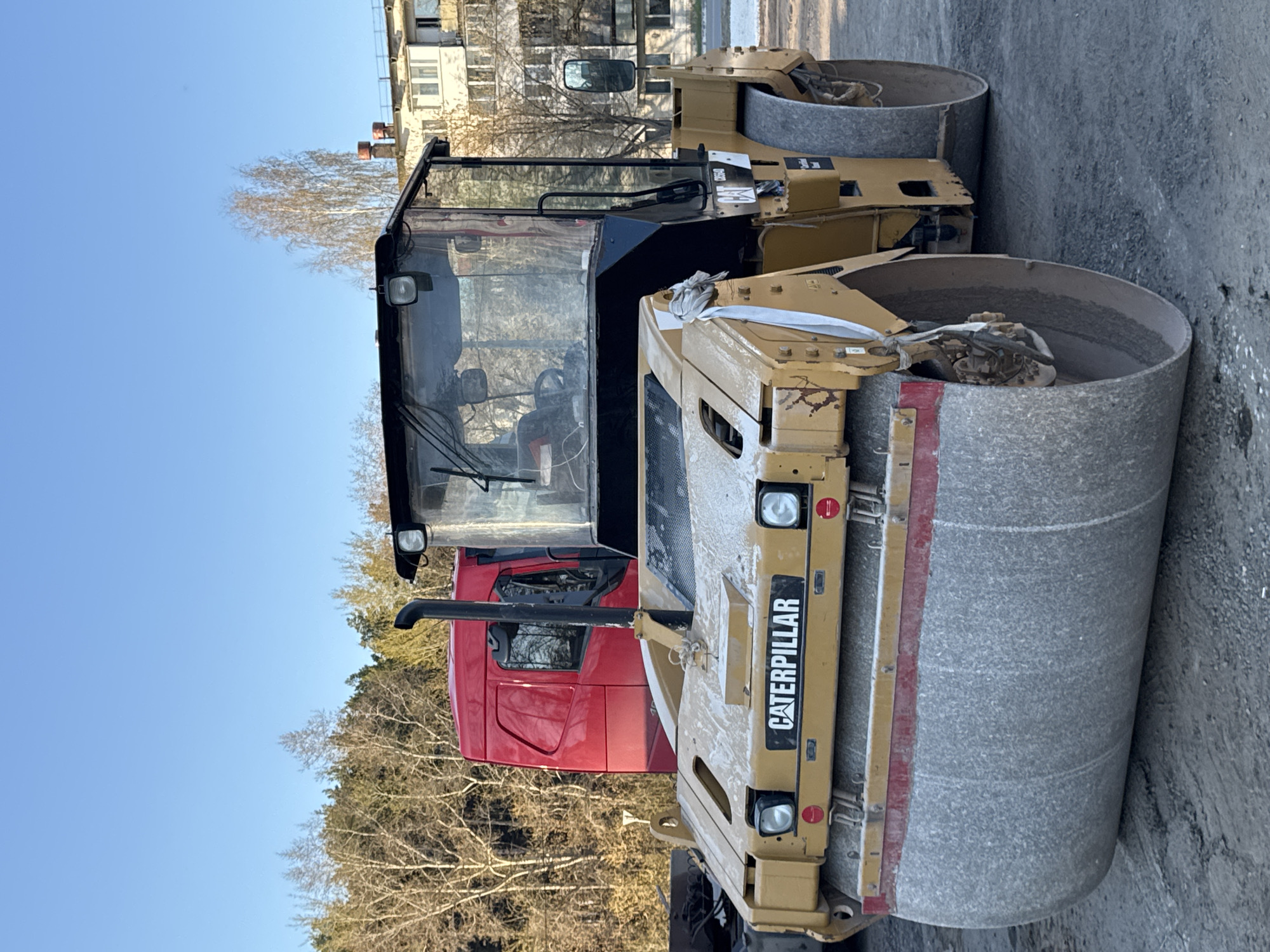 A Caterpillar road roller is parked outdoors on a paved surface. Behind the roller, the front of a red truck is visible. Trees and a light-colored apartment building are in the background. The location is likely in Chaikovsky, Russia, based on the image context.