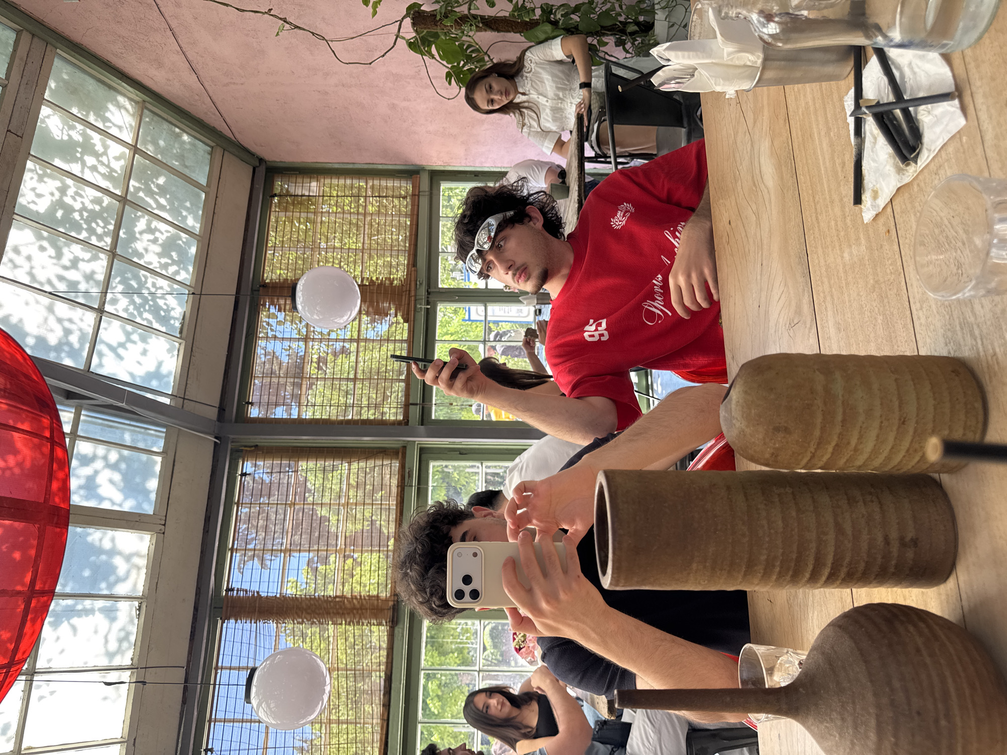 The image depicts a group of people seated at a wooden table in a brightly lit cafe with a conservatory-style ceiling. The ceiling is a mix of glass panes and wooden beams, with a large red lamp hanging from the top left. Natural light filters through the glass, casting dappled shadows of leaves onto the interior. Bamboo blinds cover the windows on the left side of the frame, revealing glimpses of greenery outside. The walls are painted a soft pink.
In the foreground, three decorative ceramic vases are arranged on the table. Two men are prominently featured. One man on the left, with curly dark hair, is holding up a smartphone and appears to be taking a photo or video. He is wearing a dark t-shirt. Another man on the right, also with curly dark hair, is wearing a red sweatshirt with "95" and "Sports Fashion" printed on it. He is looking up and to the left, holding a smartphone in his right hand. He is also wearing distinctive silver-rimmed glasses.
To the left of the foreground figures, a young woman with dark hair is seated at the table, looking towards the man taking the photo. Further back, a woman in a white shirt is seated at a table, looking towards the right. Other individuals and obscured figures are present in the background.
The overall ambiance suggests a casual dining or social gathering. The presence of bamboo blinds, the pink wall, and the specific style of the ceiling suggest a potential location in Tbilisi, Georgia, possibly at a cafe with a distinct architectural style.