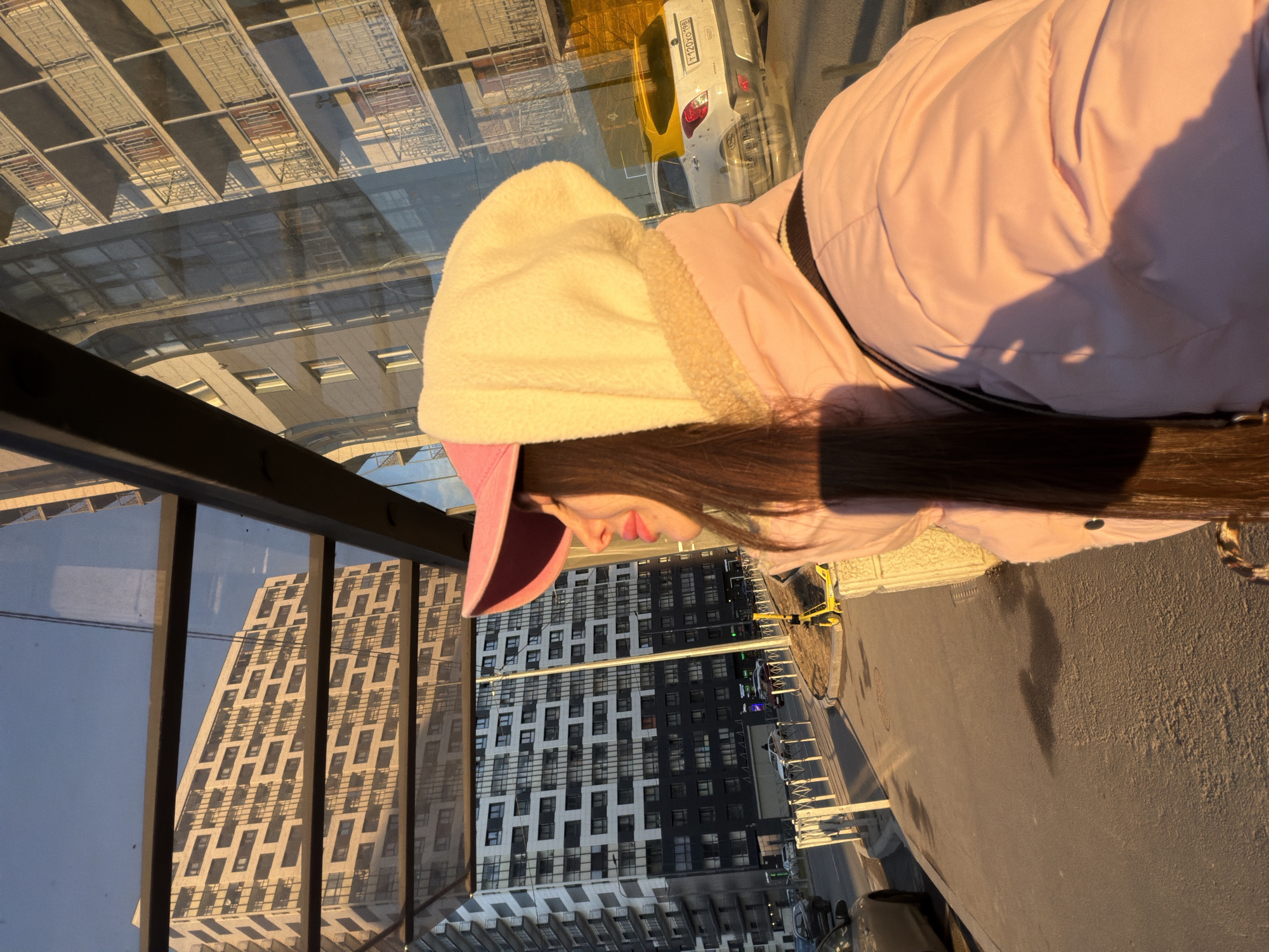 A person with long brown hair is wearing a pink hooded jacket and a pink baseball cap. The scene appears to be a bus stop or covered waiting area, as indicated by the glass panels and metal framework. Buildings, including one with a distinctive patterned facade, are visible through the glass, suggesting a city location. Cars and streetlights are also visible. The city is Saint Petersburg, Russia.