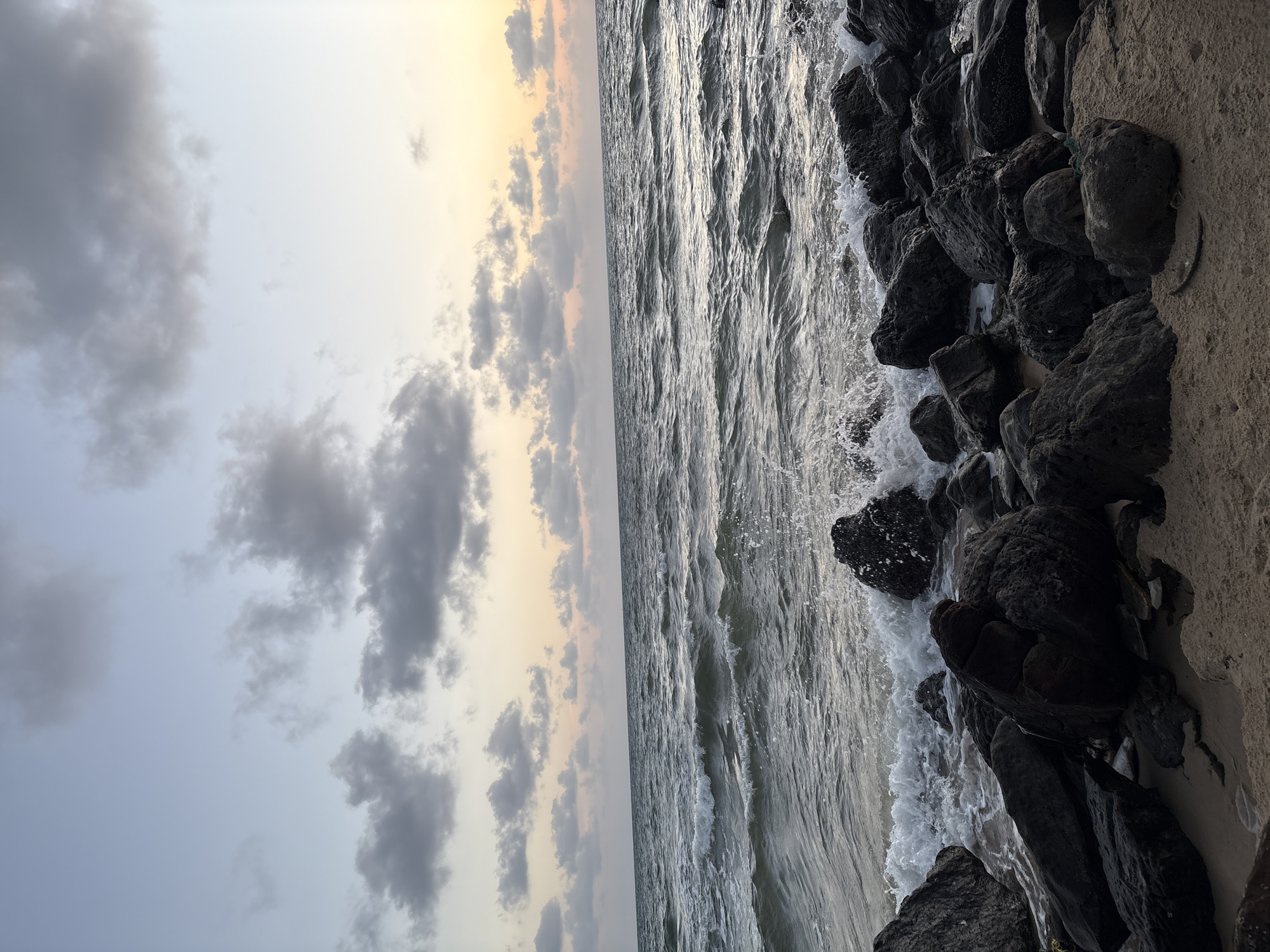 A low-angle, full shot captures a rocky shoreline meeting the ocean under a cloudy sky. The sea is a deep blue-grey, with moderate waves breaking against dark, jagged rocks. The sky transitions from a pale blue at the top to a soft yellow and peach near the horizon, where a hazy sunset is visible through scattered grey clouds. The foreground is dominated by a sandy area littered with large, dark rocks and pebbles. The scene is devoid of any people or man-made structures, giving it a natural and serene atmosphere.
