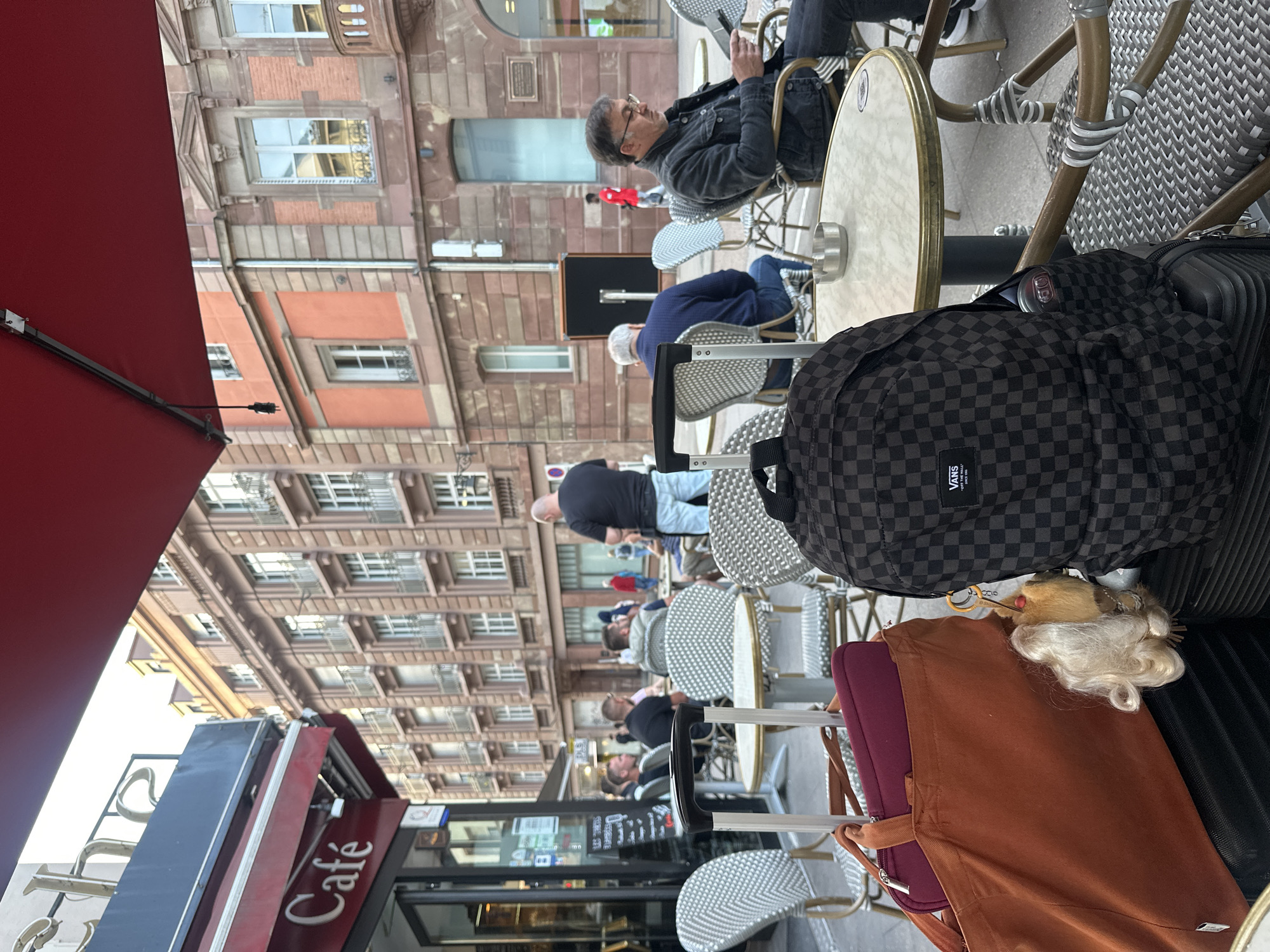 The image shows an outdoor cafe scene in Strasbourg, France, judging by the signage. Diners are seated at round tables with white wicker chairs, some holding phones and interacting. Architectural details suggest a European urban setting with multiple stories and ornate facades. A red awning is partially visible at the left, providing shade for the cafe. Luggage, including a backpack and bags, sits near some of the chairs, indicating that some individuals are likely traveling.