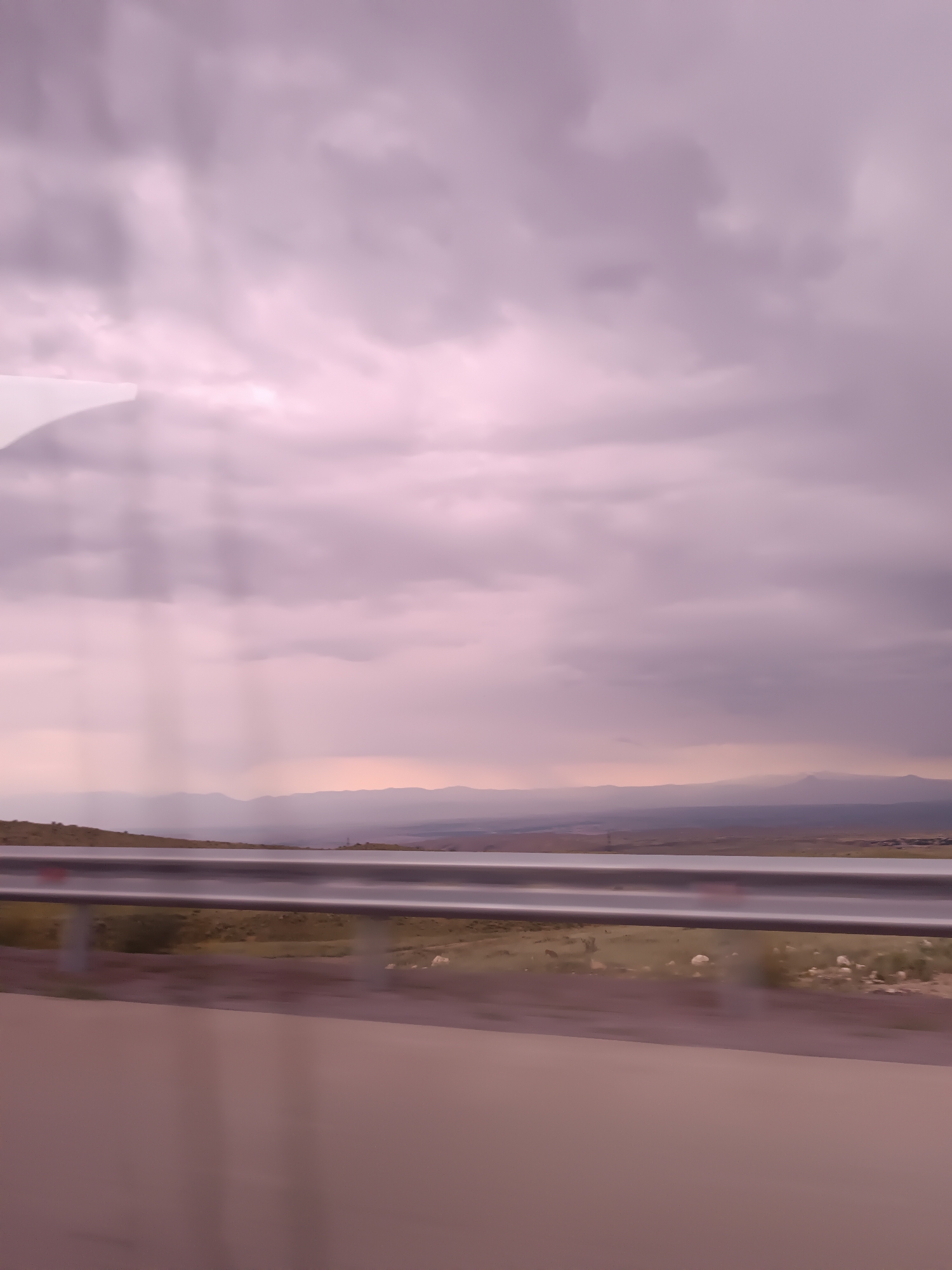 A rural landscape with mountains in the distance is visible under a cloudy sky. A guardrail runs horizontally across the middle of the frame, separating the foreground from the background. The foreground is a blurred road surface, suggesting the image was taken from a moving vehicle. The background shows a green hillside with scattered white objects that could be sheep or rocks, and a hazy mountain range in the far distance. The sky is filled with soft, muted purple clouds, with faint rays of light breaking through. The scene appears to be an overcast day in a scenic, undeveloped area.