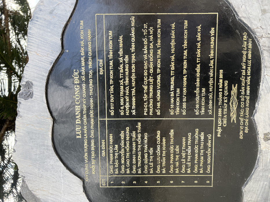 An outdoor close-up captures a dark, irregularly shaped stone plaque with a polished black surface and rough, light grey stone edges. The plaque features golden Vietnamese text, titled "LƯU DANH CÔNG ĐỨC" (Register of Merit). It identifies the project as the "Construction of Arhat Statue Garden, Kỳ Quang Pagoda" in Đăk Mar Commune, Đăk Hà District, Kon Tum Province, Vietnam. A table within the text lists nine donor families and their respective addresses, which span various Vietnamese provinces including Kon Tum, Hưng Yên, Hà Nội, and Quảng Ngãi. The inscription specifies a date of "PHẬT LỊCH 2560 - THÁNG 8 NĂM 2016" (Buddhist Calendar 2560 - August 2016) and credits "CƠ SỞ SẢN XUẤT ĐÁ MỸ NGHỆ LÂM TẠO" (Lâm Tạo Fine Arts Stone Production Facility) from Ninh Vân Craft Village, Hoa Lư, Ninh Bình, Vietnam, as the manufacturer. Blurred green foliage, consistent with tree branches, is visible in the background above the plaque, set against a bright, diffuse sky. No individuals or active interactions are present.