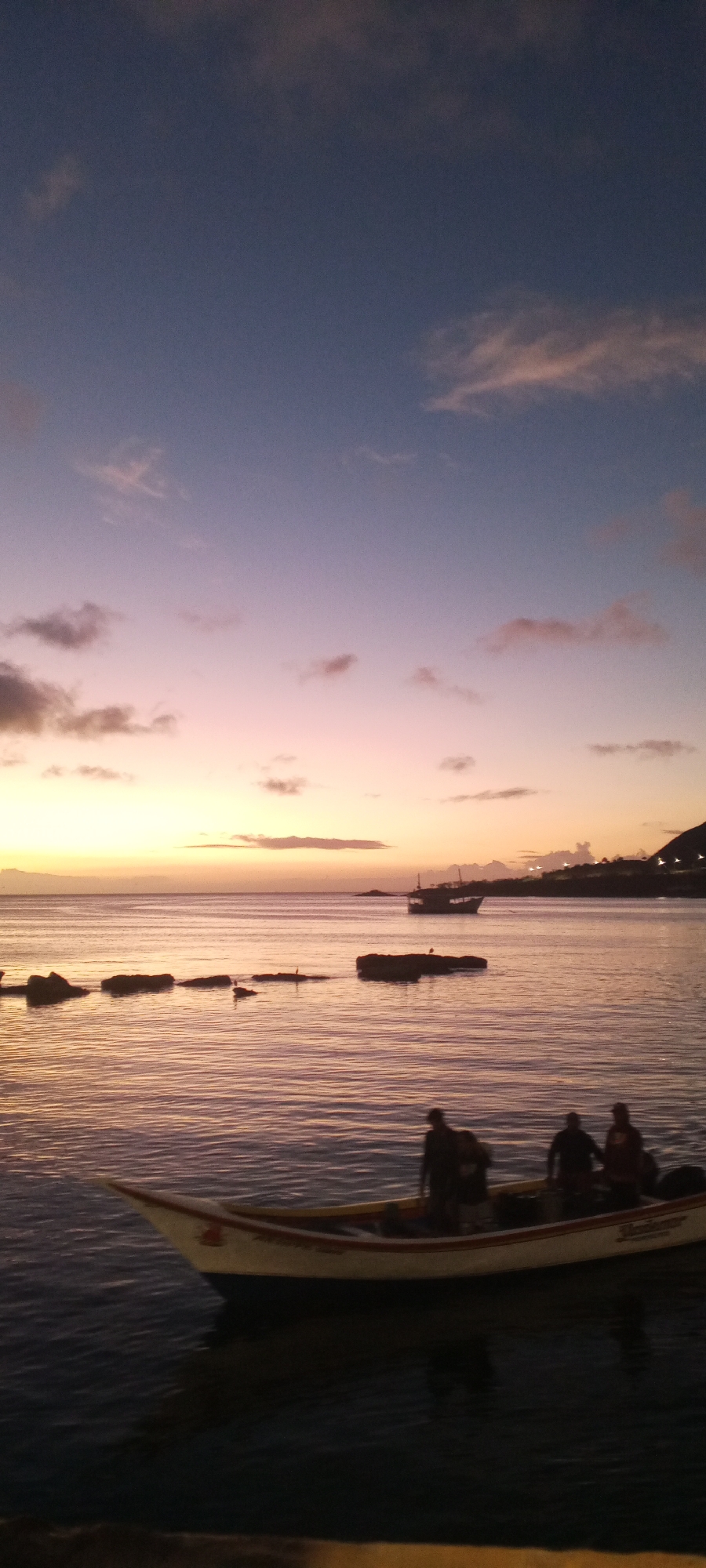 This is a vertical, full shot of the Juan Griego waterfront in Venezuela at dusk. The sky transitions from a deep blue at the top to soft purples, pinks, and oranges near the horizon, with scattered clouds. The calm water reflects the colors of the sky.
In the foreground, a white and blue boat is partially submerged at the bottom of the frame. Four dark silhouettes of people are visible standing inside this boat. The boat has a red and yellow stripe along its gunwale and some lettering on its side, though it's not entirely legible.
Further out in the water, a larger, dark silhouette of a boat with masts is sailing away from the viewer. Several dark rock formations emerge from the water in the midground, with what appear to be birds perched on some of them.
In the background, a dimly lit coastline with lights is visible, suggesting buildings and infrastructure along the shore. The overall scene depicts a peaceful evening at a coastal town.
