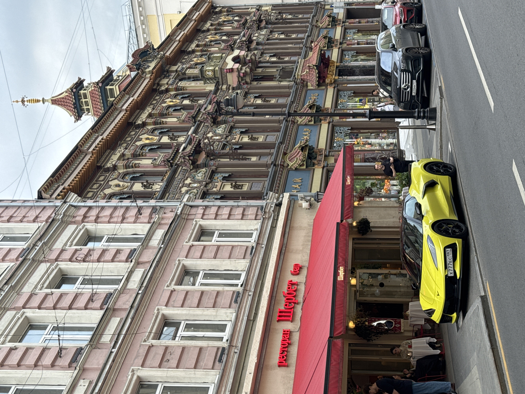 This image captures a street scene in Moscow, Russia, likely on a cloudy day. In the foreground, a bright yellow Chevrolet Corvette is parked at the curb. To the left of the car, a building with a pink facade and white trim features multiple windows. A red awning extends from the building, covering the entrance to a restaurant or bar named "Шербет" (Sherbet), indicated by red neon lettering above the awning and white lettering on the awning itself. People are seated at tables outside the establishment.
To the right of the pink building, a highly ornate building with intricate architectural details and a multi-tiered, pagoda-like roofline dominates the background. This building is adorned with colorful carvings, decorative motifs, and numerous small windows and balconies. Street lamps are visible along the sidewalk. Further down the street, a dark Mercedes-Benz GLS SUV and a red car are parked. Several individuals are present on the sidewalk, some appearing to be patrons of the restaurant and others passersby. The street is paved with asphalt, and white lines mark the parking spaces and a lane on the road. Power lines are strung overhead.