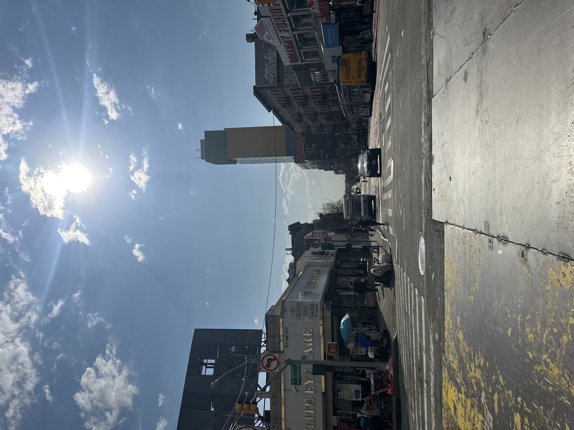 This image depicts a daytime street scene at the intersection of St. Nicholas Avenue and Juan Pablo Duarte Boulevard in Washington Heights, Manhattan, New York City, United States of America. The perspective is from the southwest corner of the intersection, looking northeast.
The bright sun, positioned in the upper right, illuminates a clear blue sky with scattered white cumulus clouds. A lens flare extends diagonally from the sun across the sky.
In the foreground, the paved street features a faded white crosswalk and a circular metal manhole cover. The immediate sidewalk area on the left shows peeling yellow paint marks.
On the left side of the street, a multi-story building houses "Elite Eye Care" at address 1422, with a prominent sign displaying a phone number "917-388-3979". Above this building, a large, dark, empty billboard structure with a visible internal frame is present. A traffic light pole with a "No Left Turn" sign and a street sign indicating "St Nicholas Ave" and "Juan Pablo Duarte Blvd" is positioned near the corner. Multiple pedestrians are visible on the sidewalk in front of "Elite Eye Care," some pushing shopping carts, and one person is partially obscured by a light blue umbrella.
The multi-lane street extends into the background. A white U-Haul truck is observed in the left lane, moving away from the viewer. A dark-colored sedan follows in a central lane. On the right side of the street, a red-painted bus lane is visible, occupied by a yellow MTA city bus (unit 3061) also moving away from the viewer.
On the right side of the street, a row of older, multi-story buildings with fire escapes and graffiti, including "ALEXANDRO," line the block. A "GALLERY FURNITURE" store sign is visible at street level. Further down the street, a tall, modern high-rise building with a distinctive green and yellow/orange facade stands prominently above the older structures. Scattered pedestrians are present on the sidewalks on both sides of the street. Utility lines and poles traverse the scene.