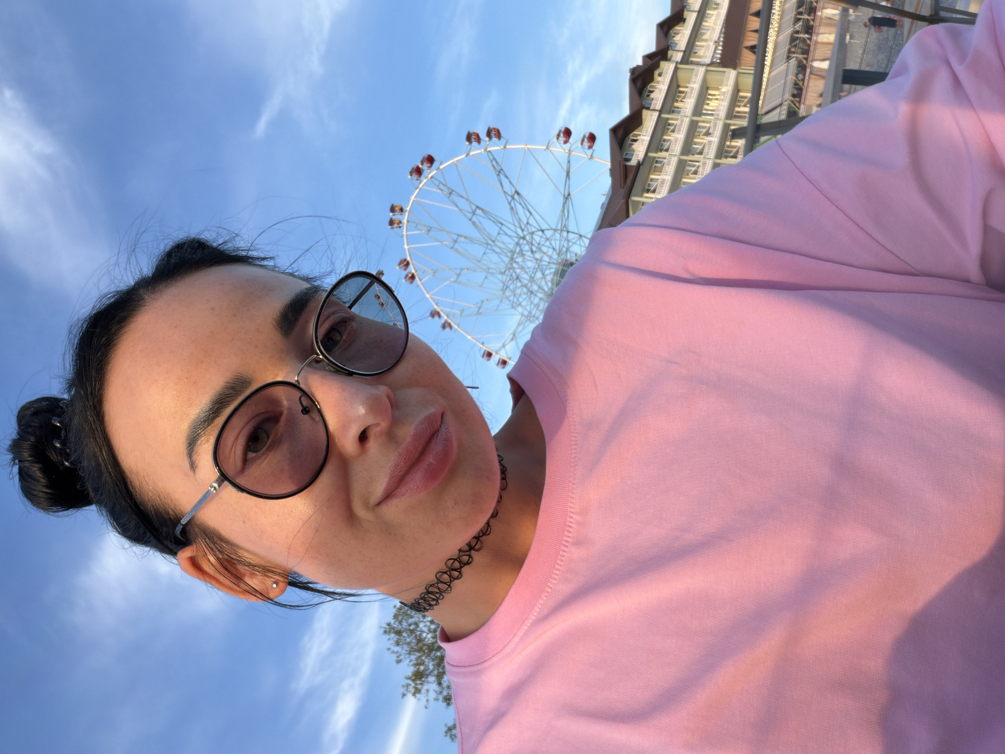 A woman with dark hair pulled into a bun, wearing round sunglasses and a pink t-shirt, is taking a selfie outdoors with a Ferris wheel and a multi-story building in the background under a bright blue sky with scattered clouds. The woman is wearing a black choker necklace. The building appears to be a hotel or resort with balconies. The Ferris wheel is white with red gondolas. The scene suggests an amusement park or entertainment area in Sochi, Russia, likely during daylight hours given the bright sunlight.