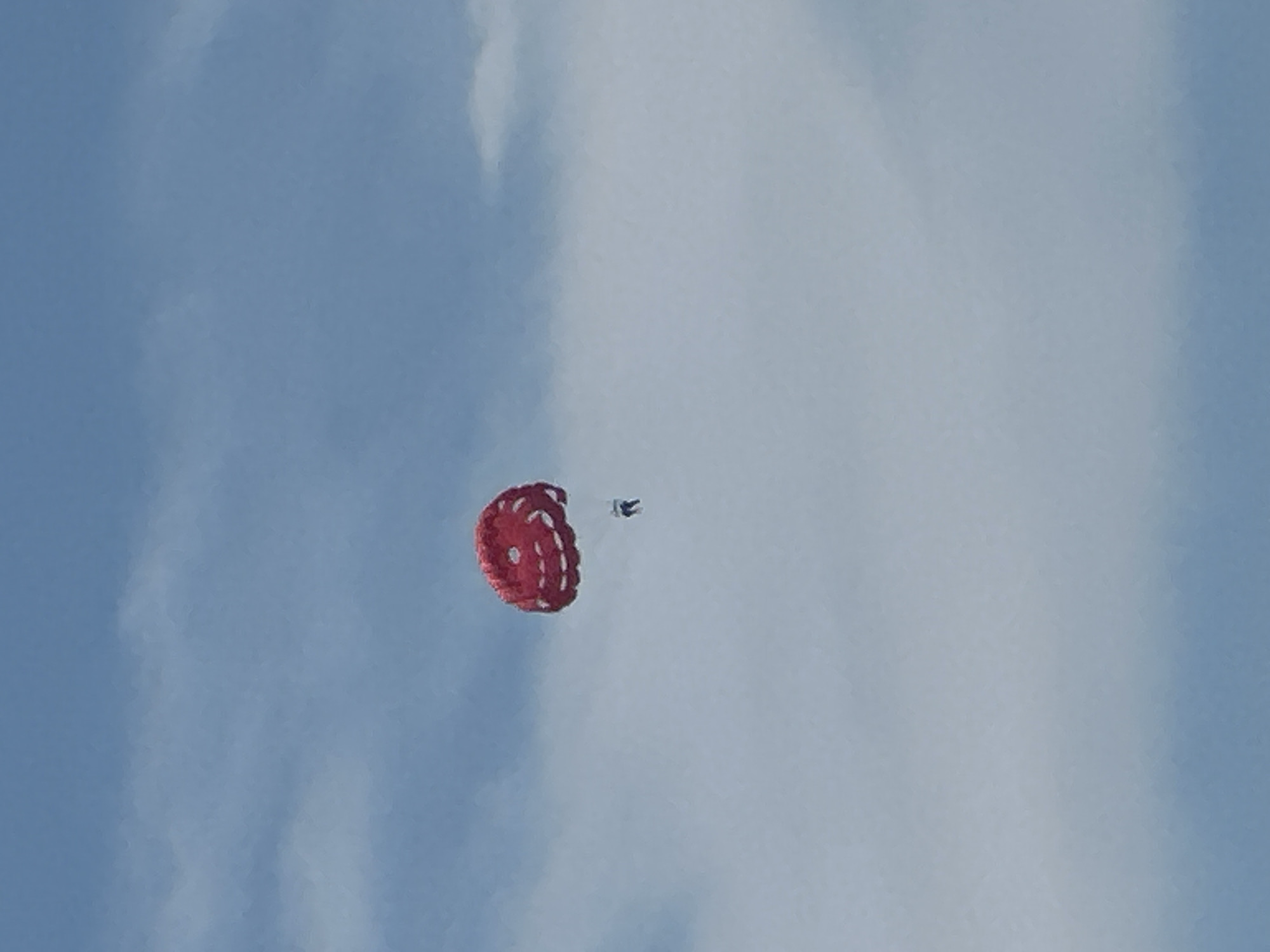 A single person is parasailing in a pale blue sky with wispy white clouds. The parasail is large and red with white circular patterns. The person is suspended below the parasail by thin ropes and appears to be wearing dark clothing. The overall impression is of a recreational activity taking place in an open, airy environment. The location of Budva, Montenegro is not discernible from the image.