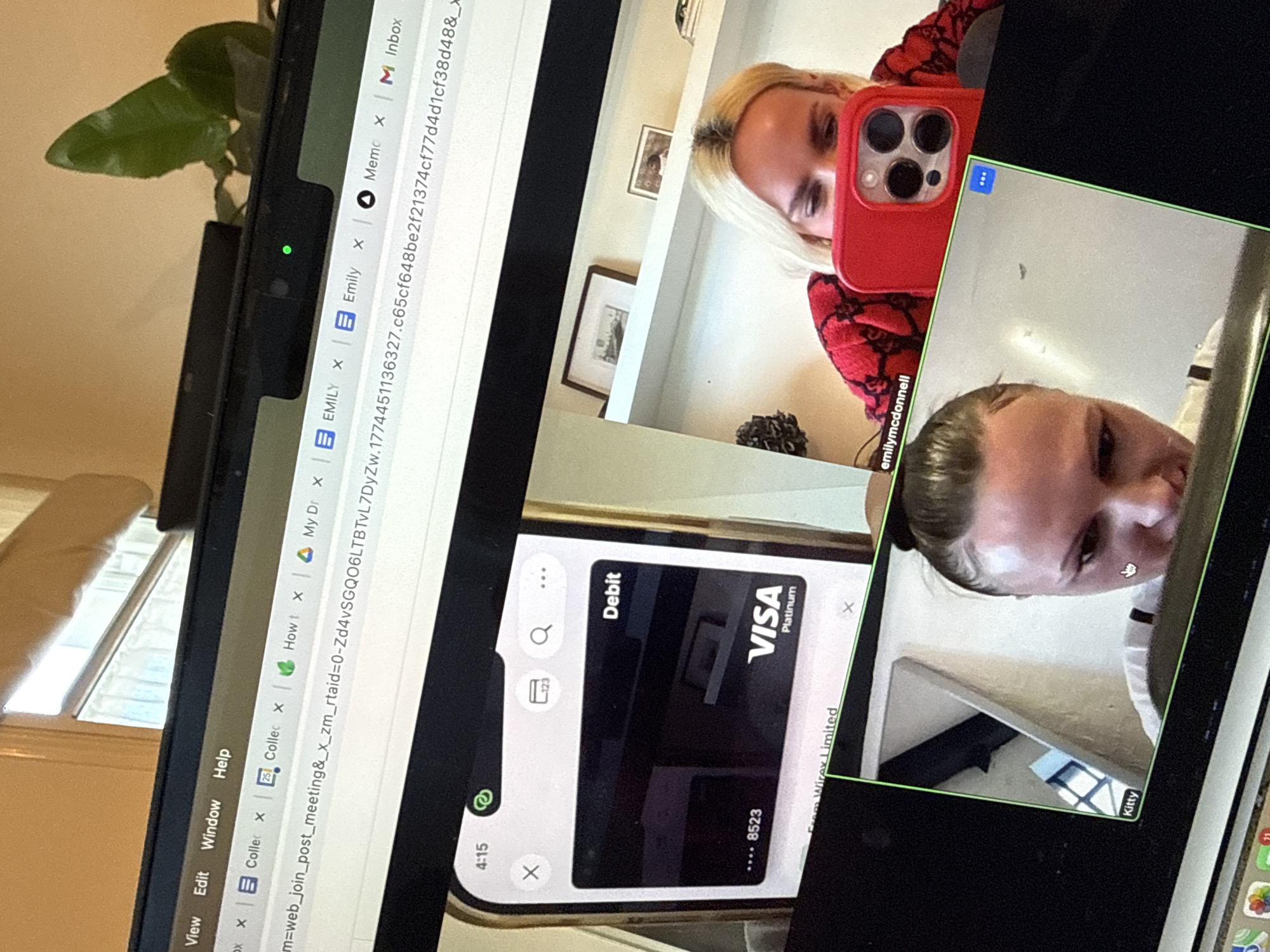 The image displays a computer monitor showing a video conferencing application alongside open web browser tabs. The scene is inferred to be related to London, United Kingdom, due to a Wirex Limited debit card being visible on a smartphone screen within one of the video feeds; Wirex is headquartered in London.
The monitor features several open browser tabs at the top, including "EMILY," "Emily," and "Inbox," indicating browsing activity. A green light on a device above the screen suggests an active webcam. Behind the monitor, part of a window and a green plant are visible.
The video conference interface shows three distinct feeds:
1. **Top-left feed:** A close-up of a smartphone screen displaying a digital "Debit" card. The card is black with "VISA Platinum" branding and shows the last four digits "...8523." "From Wirex Limited" is partially visible below the card. The phone's status bar shows "4:15."
2. **Top-right feed (labeled "emilymcdonnell"):** A woman with blonde hair and dark roots is visible, wearing a red patterned top. She holds a red smartphone with a prominent multi-lens camera, obscuring her lower face as she looks down at it. Her background includes white shelves with framed pictures.
3. **Bottom-left feed (labeled "Kitty"):** A woman with dark hair pulled back in a bun is shown leaning forward, looking down. A white mouse cursor, shaped like a hand, hovers near her face on the screen. Her background is a plain, light-colored wall.
