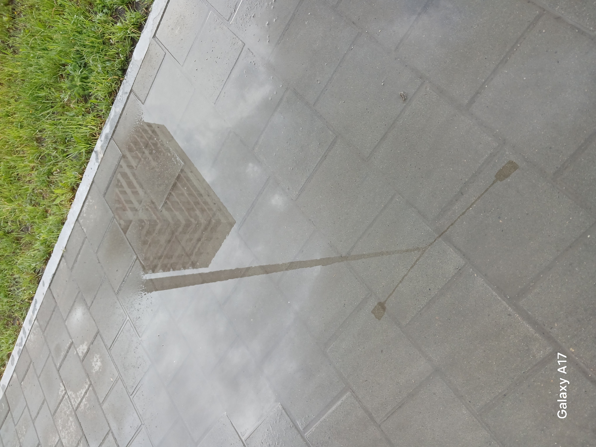 The media file displays an outdoor urban scene featuring a wet paved surface bordered by green grass. The pavement, constructed from grey rectangular or square blocks, is covered in a layer of water, creating a reflective surface. This wet pavement prominently reflects a tall, multi-story building with a grid-like window pattern, indicative of a modern or late 20th-century urban structure. Adjacent to the building's reflection, the reflection of a slender lamppost or street light is also visible.
The green grass strip runs along the left edge of the paved area, suggesting a roadside or park-like setting within a city. No people are present in the image. The overcast sky, as suggested by the reflections, indicates a cloudy day, consistent with the wet conditions. The presence of water on the pavement implies that it has recently rained or that there is standing water. The overall impression is that of a quiet moment in an urban environment in Yekaterinburg, Russia, following precipitation. In the bottom right corner, the text "Galaxy A17" is visible, indicating the device used to capture the photograph.