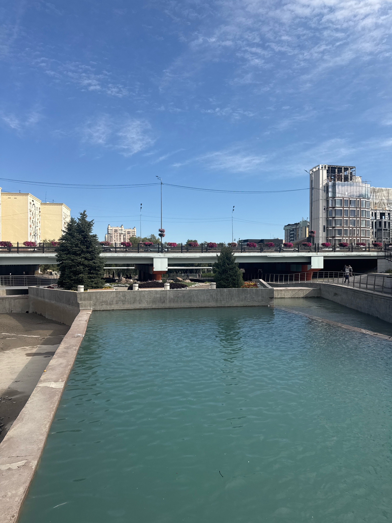 This is a photograph of a canal or river in Almaty, Kazakhstan, with a bridge and buildings in the background. The water is a greenish-blue color and appears calm. There are concrete walls surrounding the body of water. On the left side of the frame, there is a sandy embankment.
In the mid-ground, a bridge spans across the water. The bridge has a railing with decorative flower planters, featuring bright pink flowers. Below the bridge, there are visible supports and some landscaping.
On the other side of the canal, in the background, a modern glass-fronted building is under construction, with visible scaffolding and construction materials. To the left of this building, an older, lighter-colored building with many windows is visible. Further in the distance, a distinctive building with a domed roof can be seen.
Two evergreen trees are planted on the canal's edge, in front of the bridge. On the walkway on the right side of the bridge, two people are walking, one with a backpack. The sky is a clear blue with scattered white clouds. The scene suggests an urban environment with elements of nature and ongoing development.