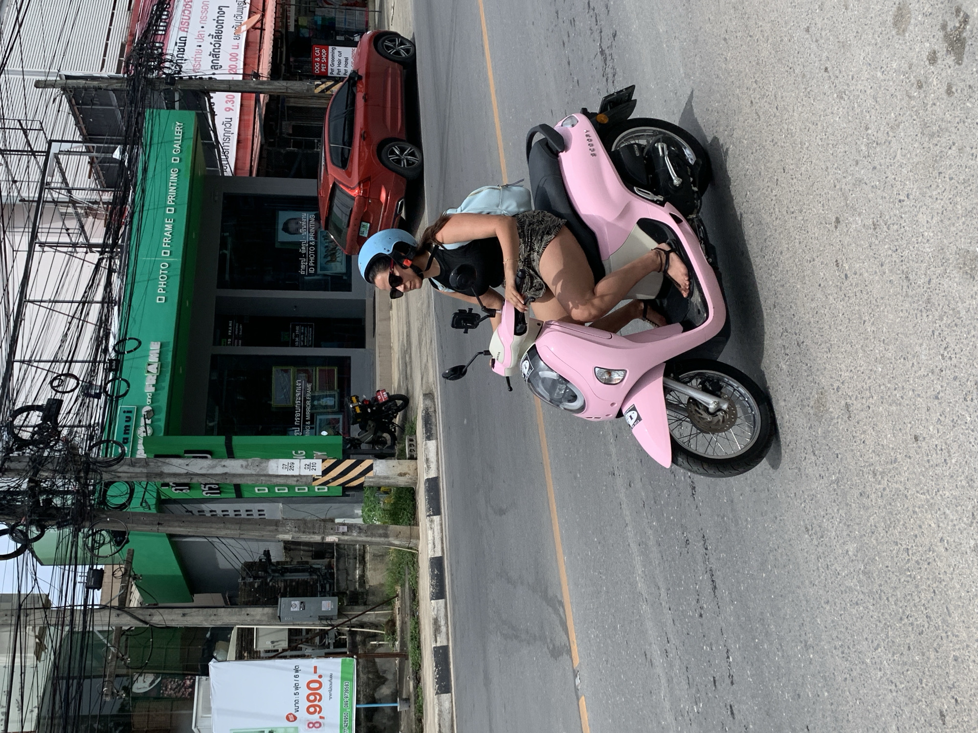 In Baan Bo Phut, Thailand, a woman wearing a light blue helmet, sunglasses, a black tank top, and a patterned skirt rides a pink scooter. She is on a paved road with a yellow dividing line. Behind her, a red SUV is parked. A green building with "PHOTO FRAME PRINTING GALLERY" signage is visible, along with a white banner displaying prices in Thai Baht. Overhead, numerous electrical wires are strung across utility poles. The scene depicts a street view with typical urban elements.