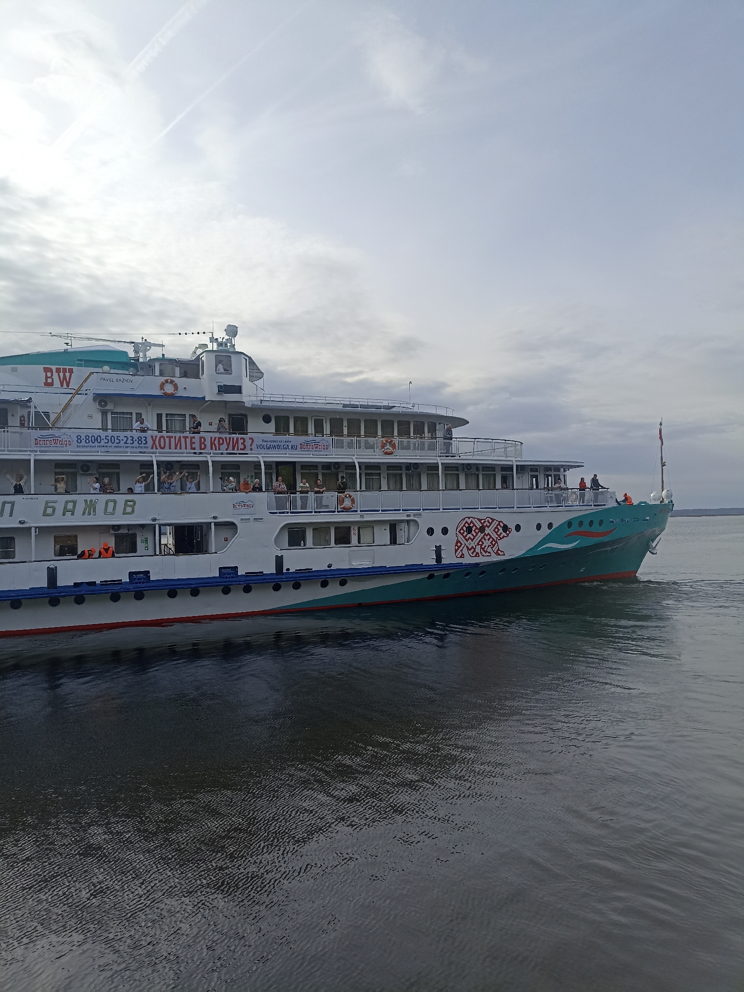 The image shows a cruise ship, named "PAVEL BAZHOV", sailing on water. The ship is white with green and blue accents and has multiple decks with people visible on them. A banner on the side advertises cruises, including a phone number and website. The water is dark and choppy, and the sky is overcast with light clouds. The location is unknown.
