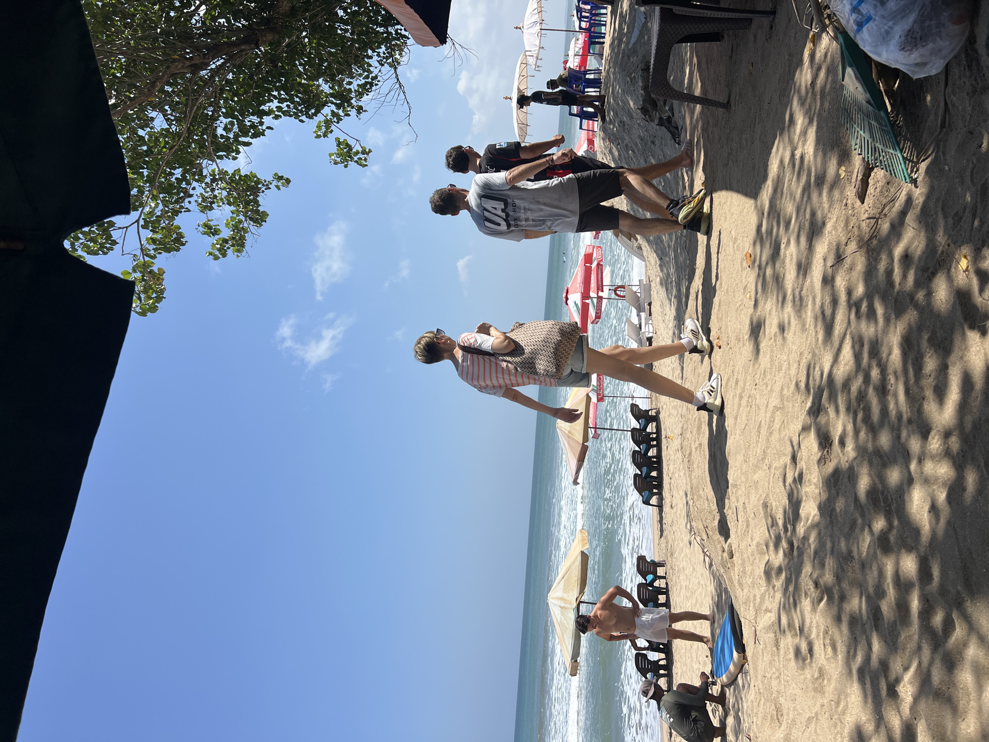 The image depicts a sunny day at Kuta Beach, Indonesia. The foreground shows sandy ground with shadows from trees, a rake, and a plastic bag. A group of people are visible on the beach and near the water. In the middle ground, several beach umbrellas and lounge chairs are set up. The ocean with waves is in the background, under a clear blue sky. Three individuals are walking away from the camera on the sand. One person is bent down near a blue and white mat. Another person is standing shirtless in white shorts. Further back, there are more umbrellas and people seated.