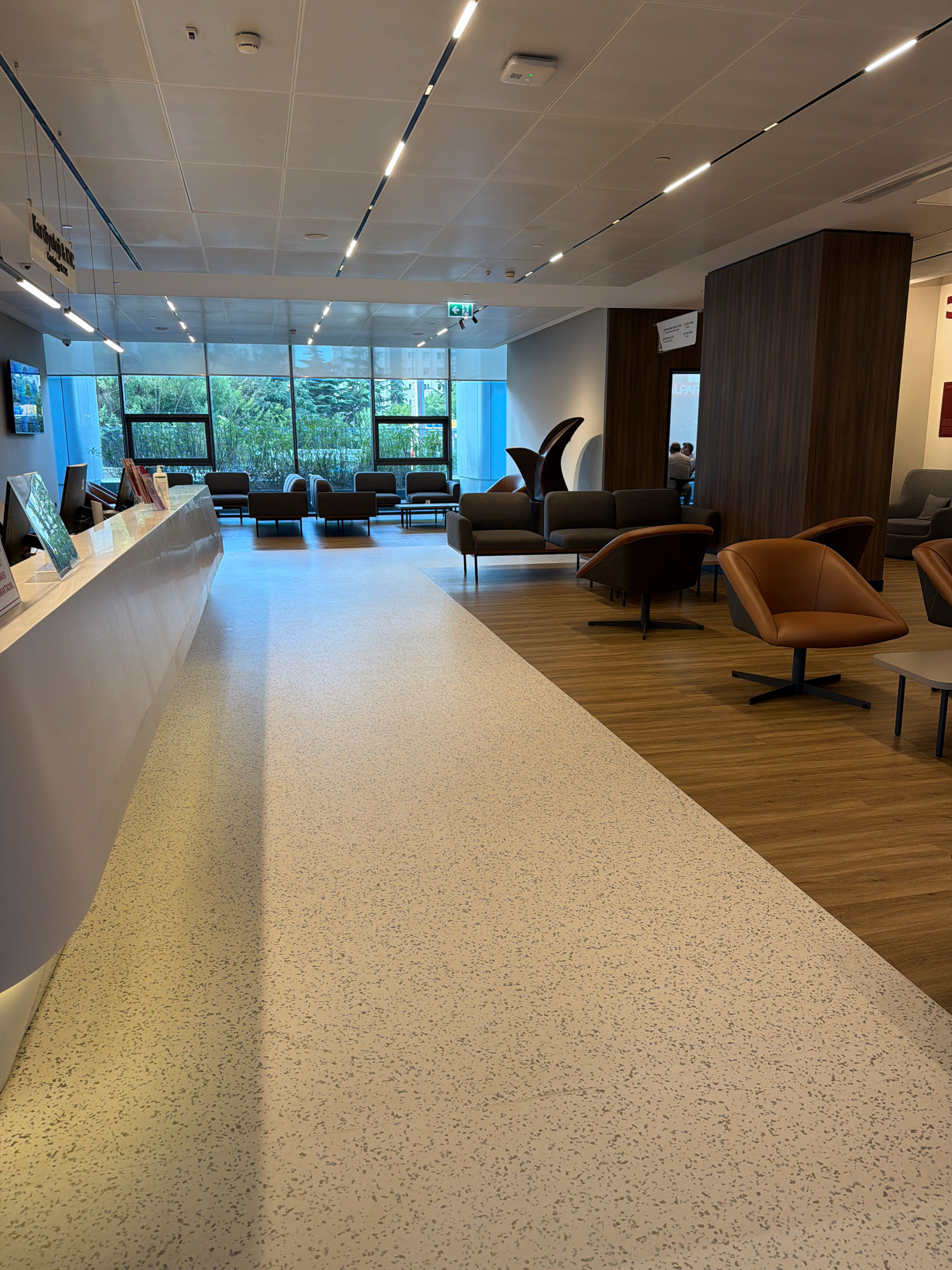 The image depicts the interior of a modern waiting area, likely a business lounge or reception area, situated in Ataşehir, Turkey. The space features a long, white, curved reception desk on the left, with a computer monitor and signage visible. Beyond the desk, large windows offer a view of lush greenery and trees. The seating area consists of several dark-colored sofas and chairs, arranged in conversation groups on a warm-toned wooden floor. In the foreground, a speckled light-colored floor leads into the seating area. A tall, dark wood-paneled pillar stands to the right, breaking up the space. In the background, a few individuals are visible seated at a table. The ceiling is modern with recessed lighting fixtures and a clean, tiled surface. The overall impression is one of a sleek, contemporary, and well-lit space designed for comfort and waiting.