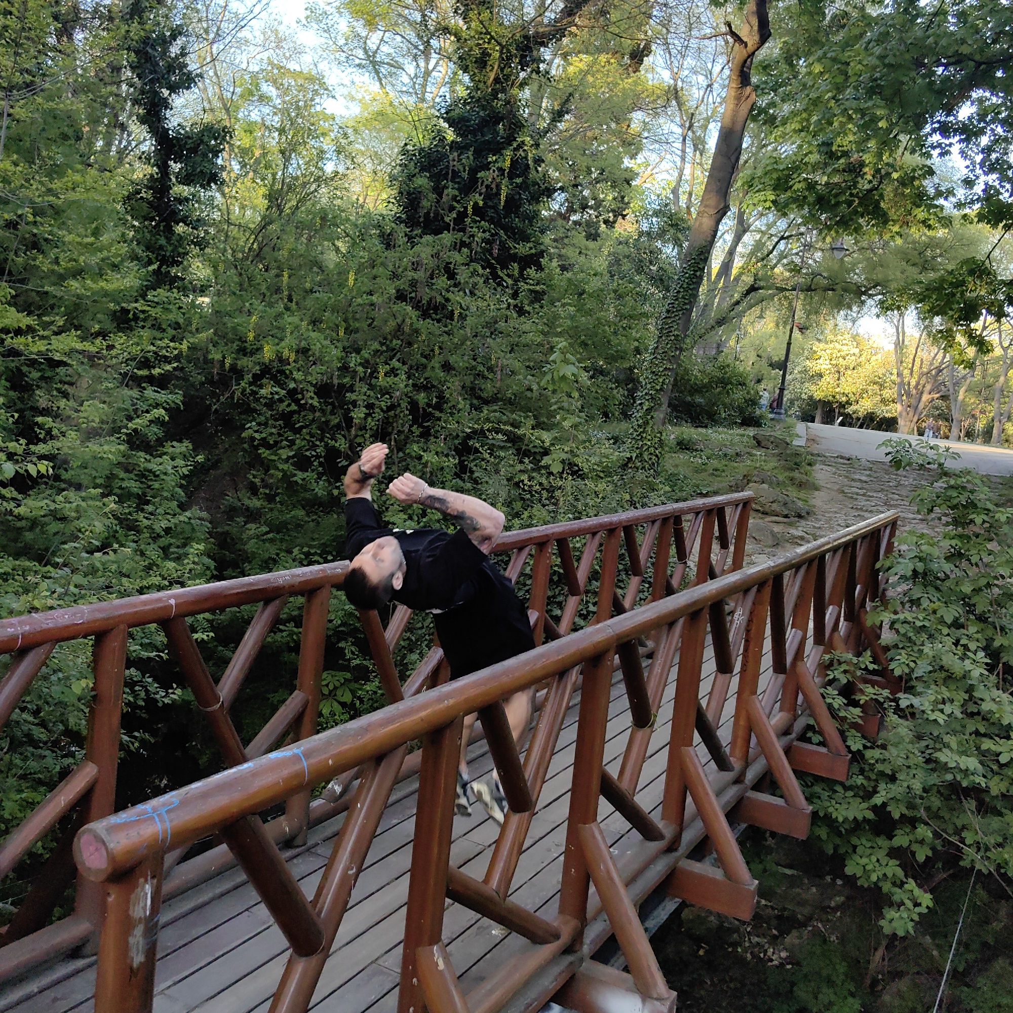A man in black clothing is leaning backwards over the wooden railing of a small bridge in a park-like environment. The bridge, constructed of dark wood, is surrounded by dense green foliage and trees. The man's arms are raised, and tattoos are visible on his left arm. The scene suggests a moment of physical activity or recreation within a natural setting, possibly a park or garden. Further into the scene, a path can be seen. Considering the available information, the location is within the city of Varna, Bulgaria.