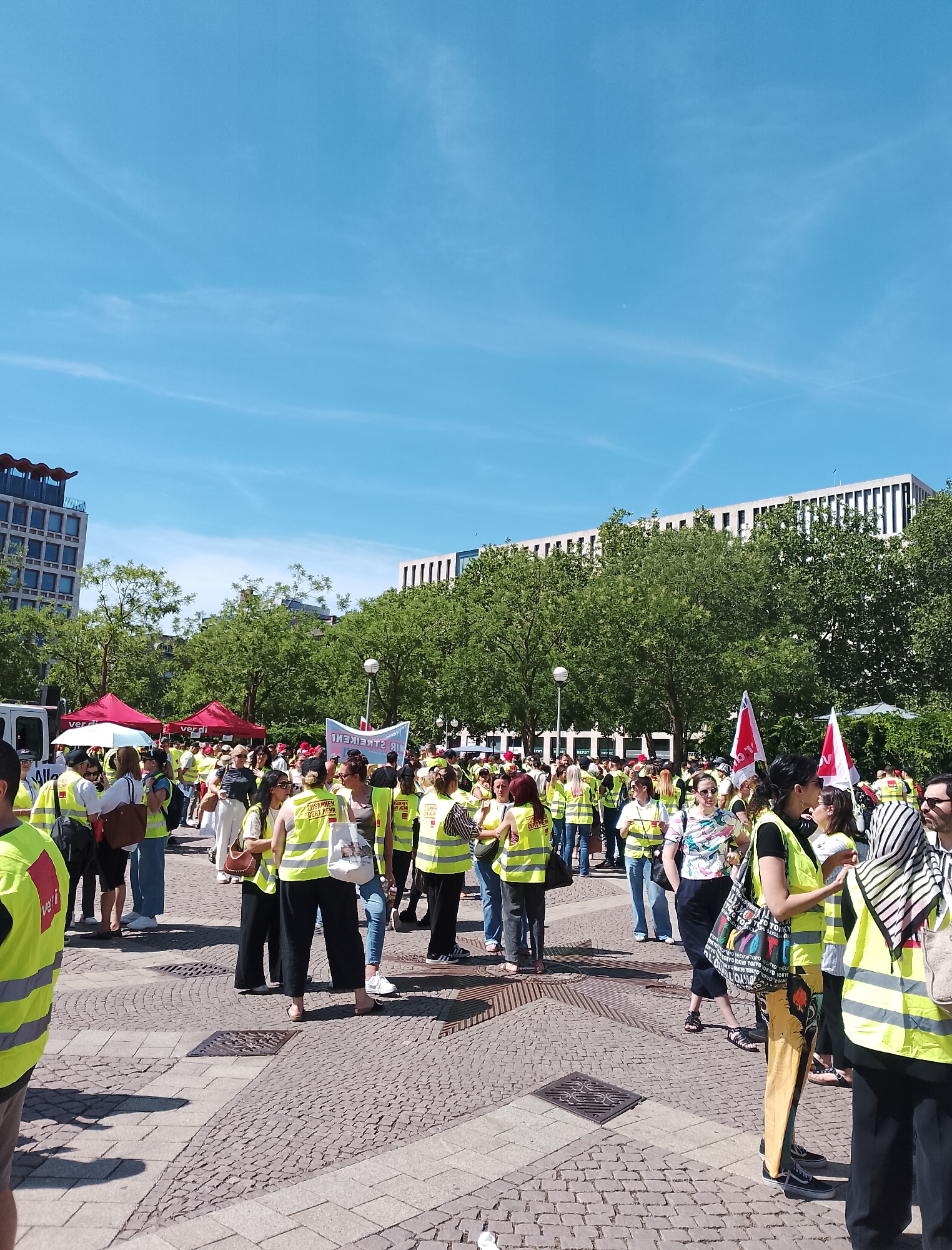 A group of people are gathered in an outdoor public square on a sunny day. The crowd is diverse, with many individuals wearing bright yellow safety vests over their clothing. Some people are holding red and white flags, one of which bears text in German. In the background, there are trees and modern buildings, suggesting an urban environment. Two red tents are set up, one with the logo "ver.di" visible. The ground is paved with interlocking stones and several metal grates are present. The sky is clear and blue with some wispy white clouds. The overall impression is that of a protest or a rally.