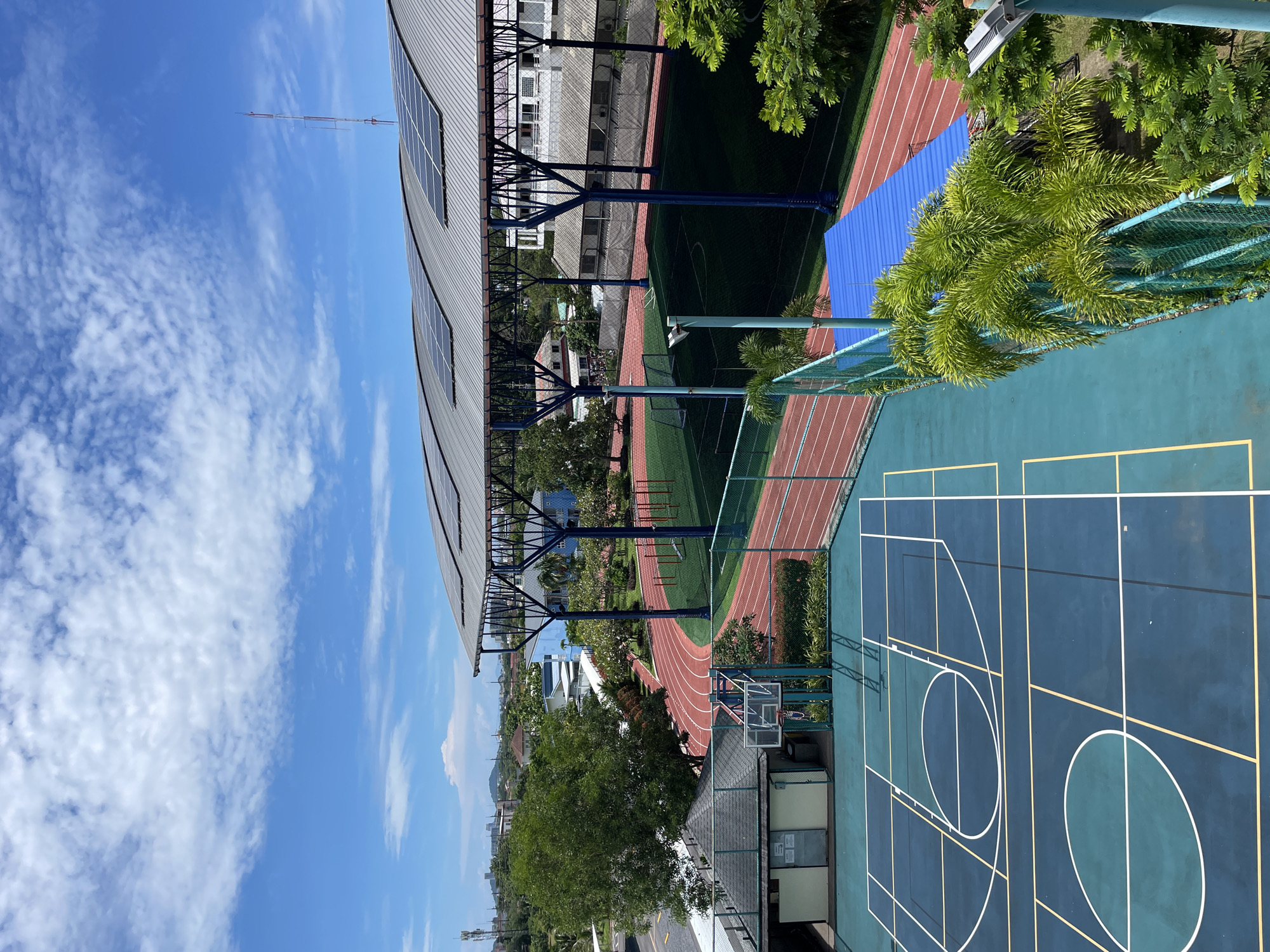 This high-angle outdoor photograph captures a sports complex on a sunny day with a bright blue sky filled with scattered white clouds. In the foreground, a teal-colored basketball court is marked with white and yellow lines. Adjacent to it, a small, dark-roofed building with a basketball hoop stands. Beyond the basketball court, a red running track encircles a green artificial turf field, which appears to be a soccer field with visible goals. A large, gray, arched roof structure with solar panels spans over part of the sports facilities. In the background, trees, buildings, and a communication tower are visible, suggesting an urban or suburban setting. The scene is an unobstructed view of a well-maintained athletic facility.