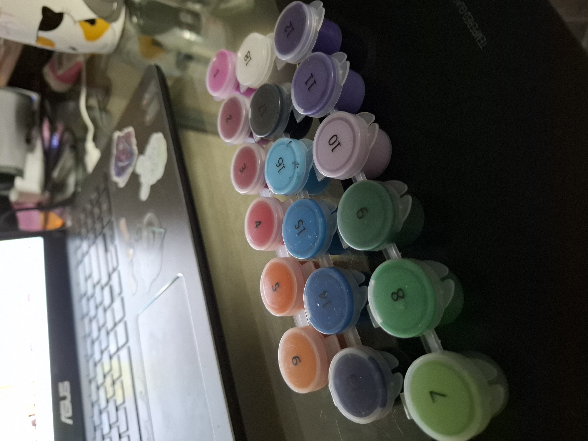 The image presents a close-up, oblique overhead shot featuring a row of small, interconnected plastic paint containers, typically used for paint-by-number kits. The containers, arranged in a diagonal line from the bottom right to the top left, are filled with various colors, each labeled with a number. From the bottom to the top, the paints are in the following colors: green, dark blue, orange, blue, orange, red, light blue, red, light blue, grey, pink, white, purple and pink.
To the left of the paint containers is part of a laptop. Stickers are visible on the laptop's case.
A black surface is visible adjacent to the containers and a laptop. It is unclear where the image was taken.
