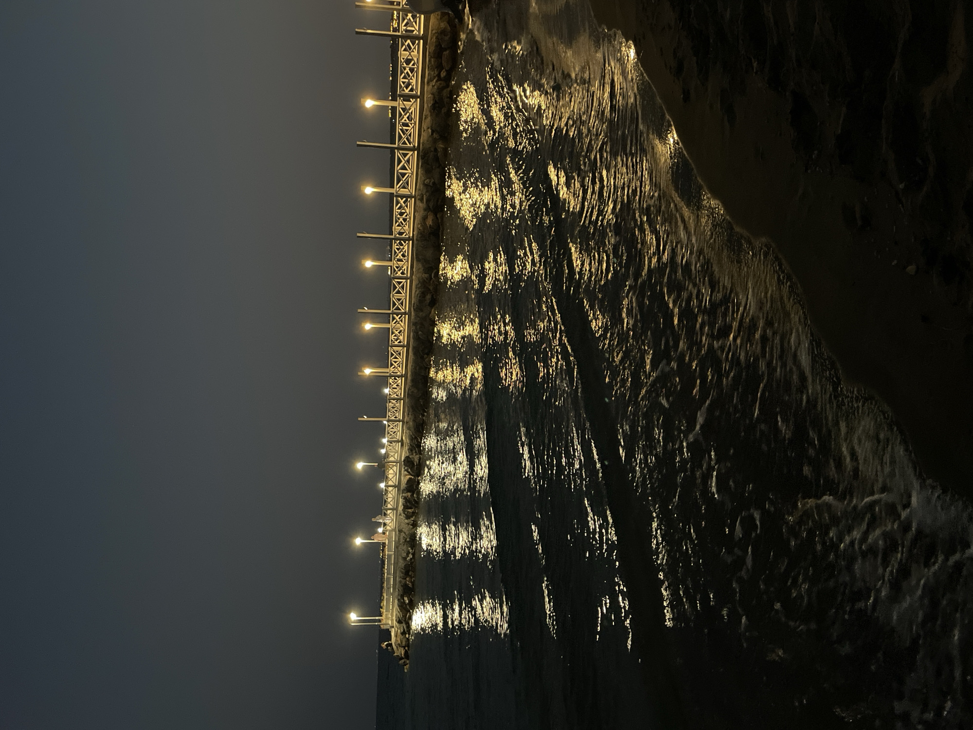 The image depicts a pier at night in Trikomo, Northern Cyprus. The pier is illuminated by a series of lights, casting a warm glow onto the water below. The water is relatively calm, with gentle waves lapping against the shore. There are a few people visible on the pier, appearing to be enjoying the evening ambiance. The sky is dark, indicating it is nighttime. The overall scene conveys a sense of tranquility and peace.