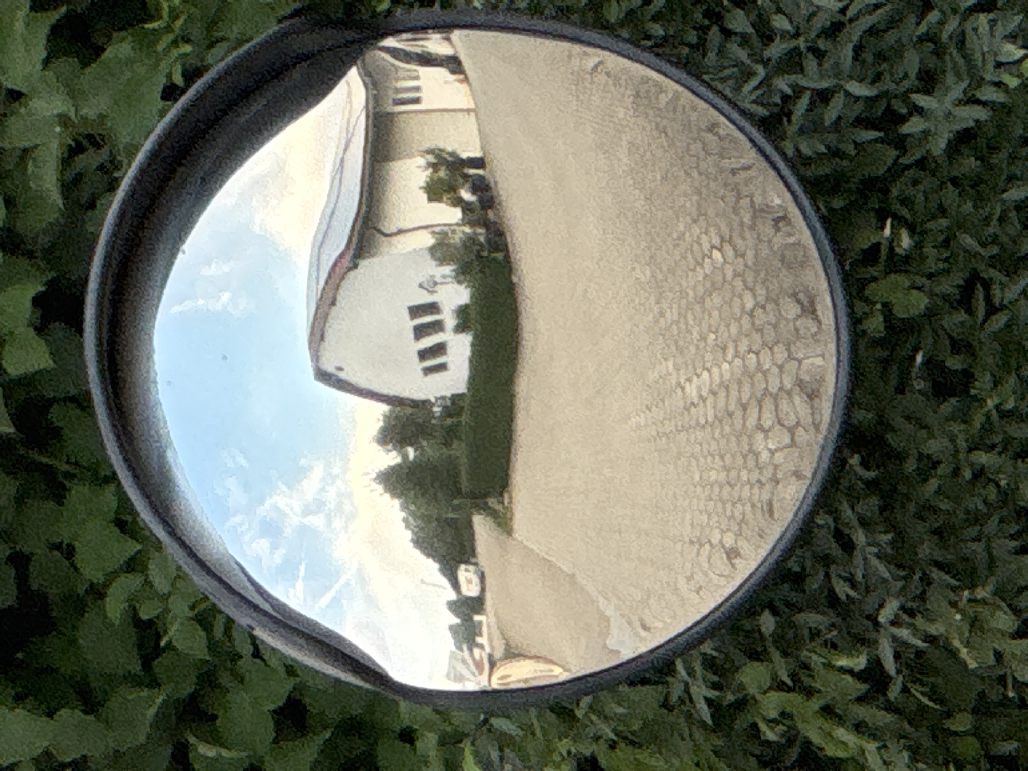 A convex mirror, mounted outdoors and surrounded by lush green foliage, reflects a scene of a paved driveway leading to a building. The building, with a light-colored facade and a curved, tiled roof possibly equipped with solar panels, features several windows. To the left of the building, a white caravan or RV is visible on the driveway, parked near some trees and a hedgerow. The sky above is partly cloudy. The image captures a wide-angle view, characteristic of a convex mirror, distorting the proportions and expanding the field of vision. The setting appears to be a rural or suburban area, possibly a residential or commercial property.