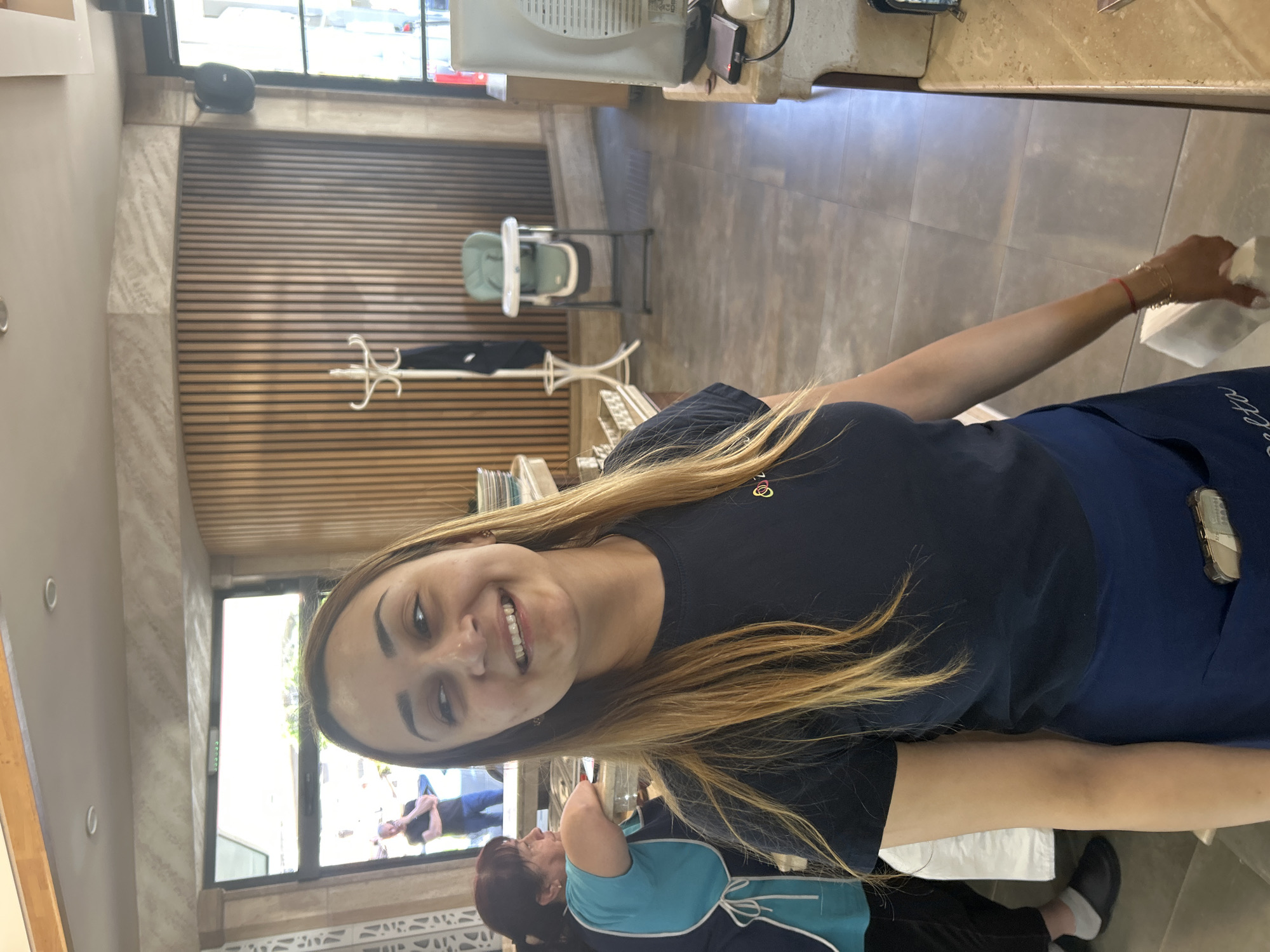 A medium shot depicts a smiling young woman in a navy blue t-shirt and blue apron. She has long blonde hair. She is holding a white object in her right hand. Behind her, a woman is partially visible in a blue and teal uniform. A window reveals people outside on a street. The interior includes a light-colored wooden wall, a white coat rack, a high chair, and beige tile flooring. The location is Kazanlak, Bulgaria.