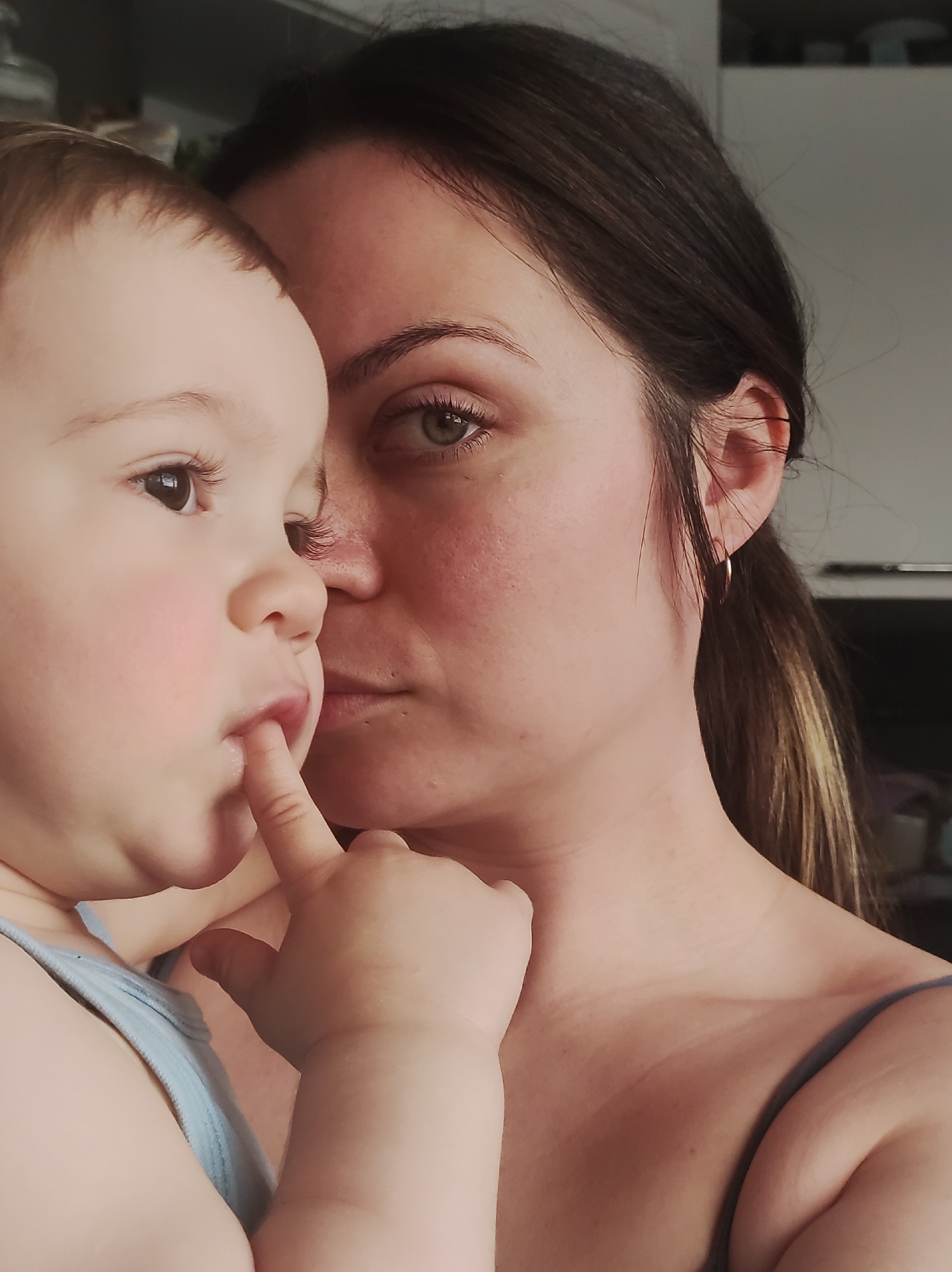 A close-up, intimate portrait captures a woman holding a young child, likely a mother and her baby or toddler, in an indoor setting in Chaikovsky, Russia. The woman, positioned on the right, looks towards the right side of the frame with a serious or pensive expression. She has dark hair tied back, striking green or hazel eyes, and a small golden hoop earring in her visible right ear. She is wearing a light-colored strap top.
The child, on the left, is partially visible from the forehead down, facing away from the camera with its face mostly in profile. The child has light brown hair and prominent rosy cheeks. A tender moment is captured as the child has a finger playfully or thoughtfully placed in its mouth. The child wears a light blue strap top. The connection between the two subjects is highlighted by their close proximity and the soft, diffused lighting. The background is out of focus, suggesting a domestic environment. There is no visible text in the image.