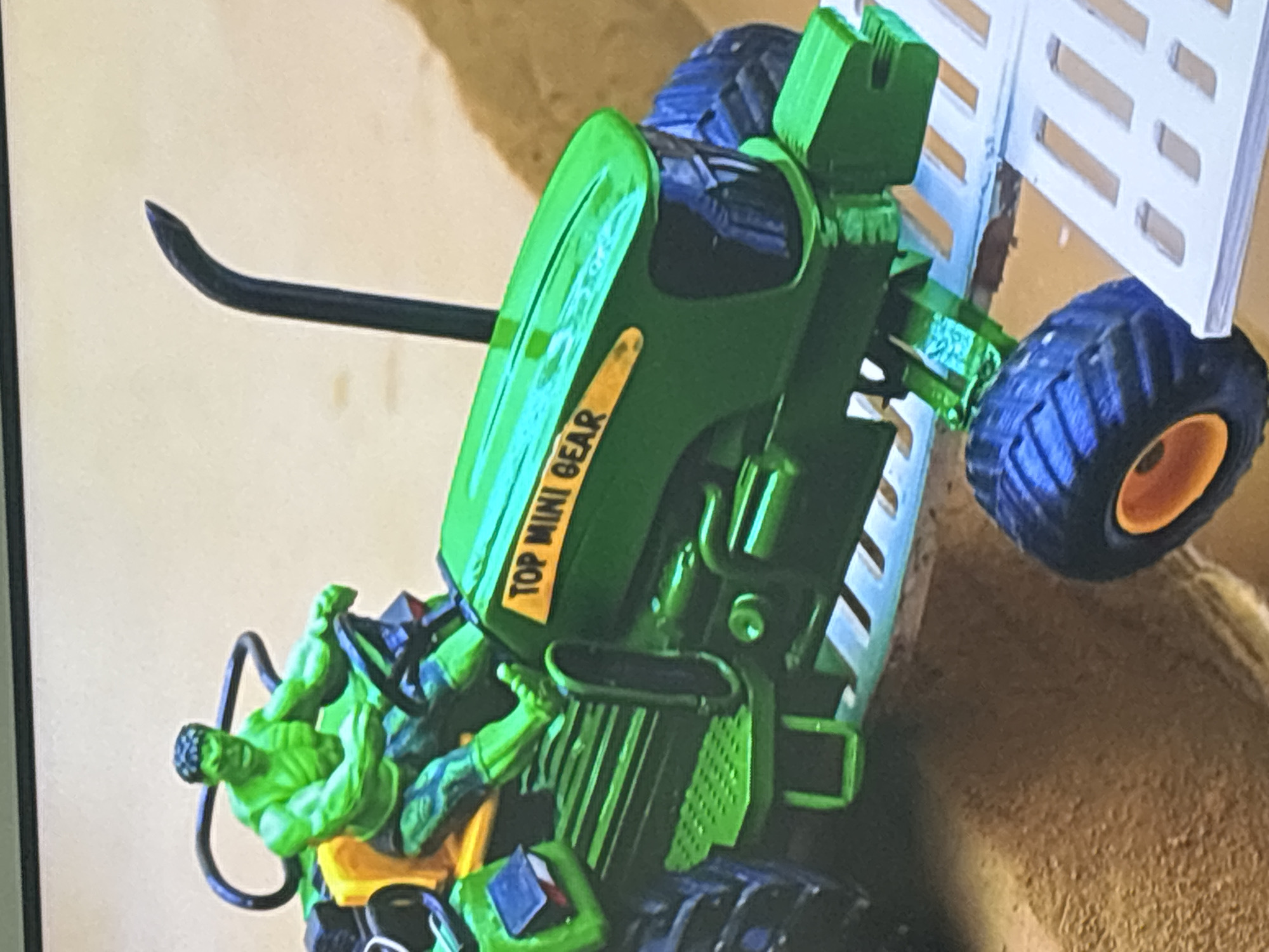 A close-up shot shows a green toy tractor with a green Hulk figurine driving it. The tractor has "TOP MINI GEAR" written in yellow on its hood. The Hulk is seated at the steering wheel, appearing to be in control of the vehicle. The tractor is positioned on a sandy surface and a white ramp-like structure. The background is a plain, light-colored surface. There is no indication of the city of Dubai, United Arab Emirates, in this image.