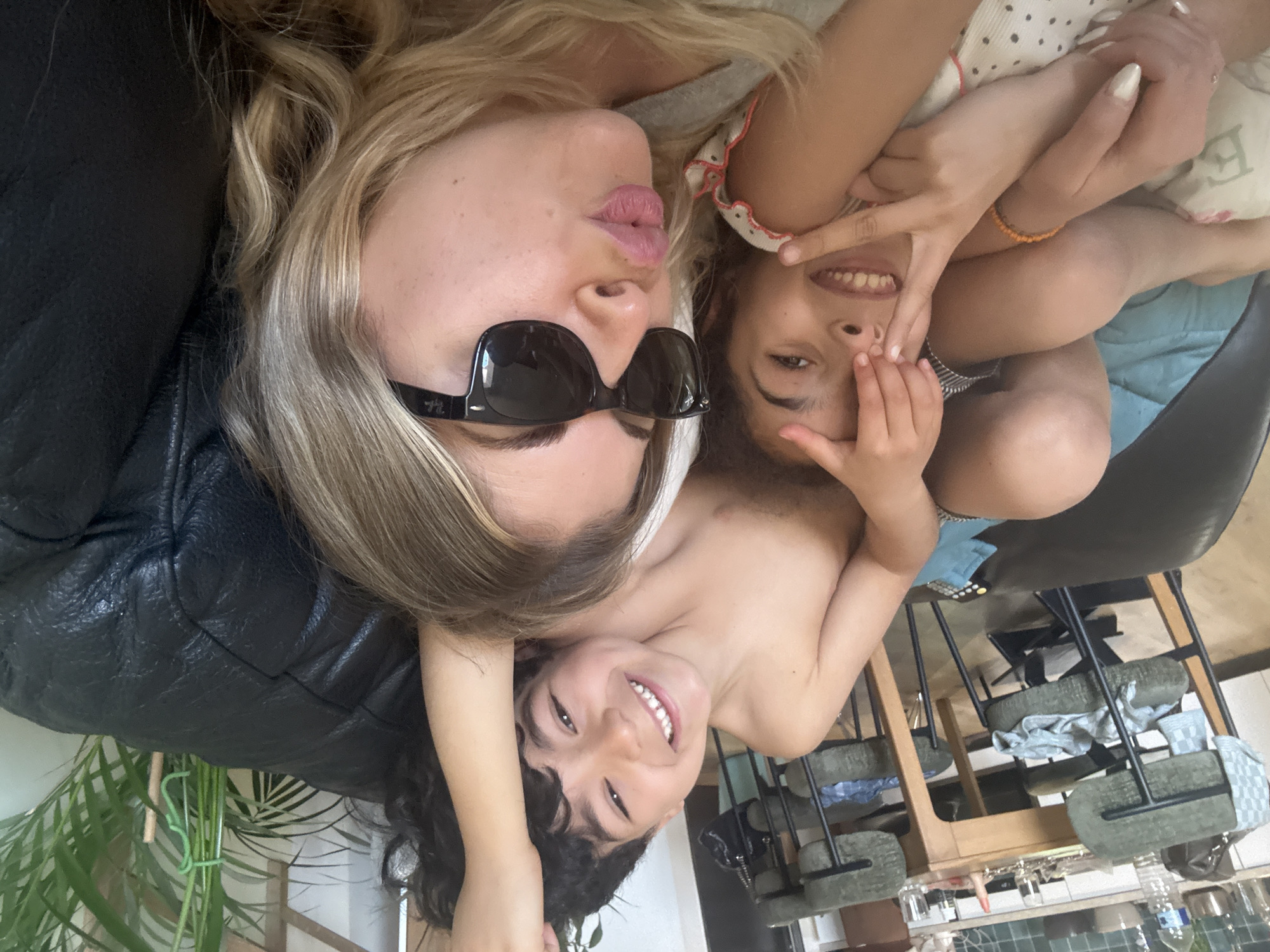This image captures an adult woman and two children playfully posing for a photo in an indoor setting, taken from an overhead perspective while they lie down. The original image is rotated 90 degrees counter-clockwise; the description assumes a correct upright orientation.
In the upper left, an adult woman with long blonde hair lies on a black leather-like surface, possibly a couch. She wears black sunglasses, with a faint 'Ray-Ban' logo visible on the arm, and makes a playful kissing face with pursed lips. To her right, a child with dark, curly hair smiles broadly, playfully covering part of their face with their hands. Another child with dark hair also smiles broadly in the lower center, looking upwards. An adult hand with painted nails and a beaded bracelet is visible near the first child.
The background depicts a domestic scene. A green houseplant is visible on the left. To the right, a dining or kitchen area features wooden tables and chairs with green fabric seats and metal frames. A blue cloth and a remote control rest on a surface. Further back, a tiled kitchen wall displays hanging glasses and a water bottle. Faint, unclear text is visible on a pillow held by one of the children.
The atmosphere is joyful and affectionate, suggesting a happy, playful interaction during the daytime. While the context places the image in Rousset, France, no specific visual cues within the image confirm this location.