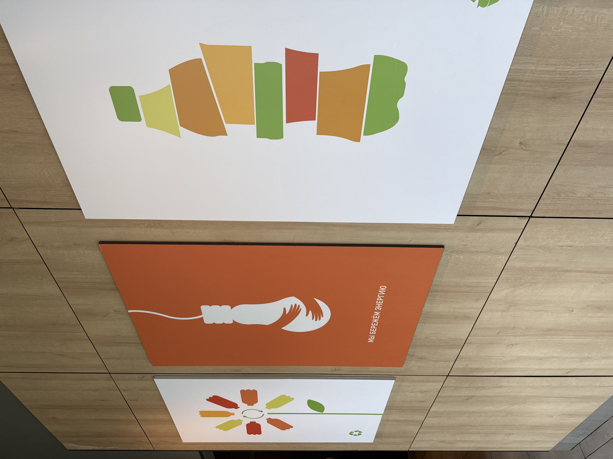 The image displays three posters on a light-colored wooden wall. The posters are arranged horizontally. The leftmost poster is white with a graphic of a flower made of recycled plastic bottles, with a green leaf and stem, and a recycling symbol at the bottom. The middle poster is orange with a white graphic of a lightbulb being embraced by two hands, and text in Russian below that translates to "We save energy." The rightmost poster is white with a graphic of a bottle or a stylized figure made of colorful geometric shapes in shades of green, yellow, and orange. The posters are mounted on a wall paneled with wood grain. The overall setting appears to be indoors, possibly in a public space or educational institution, suggesting a theme of environmental awareness and energy conservation.