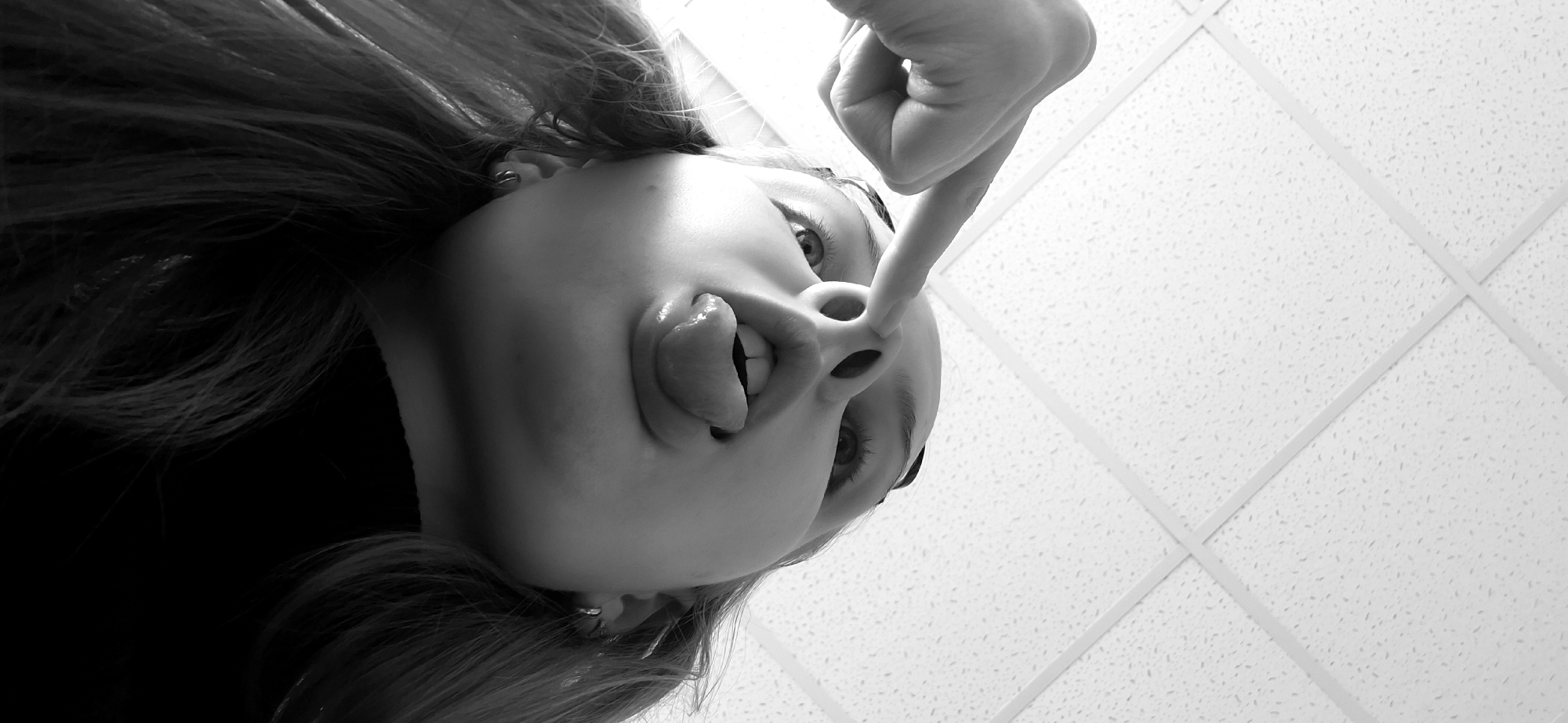 A black and white, low-angle photograph depicts a person's inverted head. The individual, with light-colored hair cascading to the sides, appears to be looking downward towards the camera. The left ear is visible, adorned with multiple hoop earrings. The person's tongue is extended upwards from the mouth, making contact with the nasal area, and the upper lip is raised, revealing the upper teeth. The index finger of a left hand is inserted into the person's right nostril, causing visible distension. The background consists of a white acoustic tile ceiling with a visible grid pattern and speckled texture, characteristic of an indoor environment. The specific location within the city of Chaikovsky, Russia, cannot be determined from the visual information presented in the image.