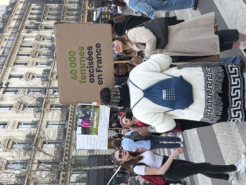 A street-level photograph captures a public demonstration in Paris, France, set against a backdrop of classic multi-story Haussmannian buildings featuring ornate facades and wrought-iron balconies. The scene takes place on a paved surface, likely a city street or square, with faint crosswalk markings visible. Bare trees line the street, and a "McDonald's" sign is visible in the upper right background.
In the foreground, a person with their back to the camera wears a cream-colored, long-sleeved garment with a black and white geometric pattern and fringe along the lower edge, a dark head covering, and a blue drawstring backpack with a white "TBS" logo. To their immediate left (viewer's right), a woman in a light beige trench coat holds the bottom left corner of a large cardboard sign and carries a dark shoulder bag. Her gaze is directed upwards and to the left. A third woman, partially obscured between the first two, holds a smartphone.
The large cardboard sign prominently displays text in green and purple, stating: "60 000 femmes excisées en France" (60,000 women excised in France). Further back and to the left, another sign is visible, featuring a photograph of a woman and the name "Gisèle Pelicot," accompanied by handwritten French text beginning "Après tant de..." (After so much...).
To the far left of the central figure, a woman wearing a white t-shirt, black fitted trousers, white sneakers with green accents, and sunglasses holds a red backpack and looks towards the left of the frame with her mouth slightly ajar. Numerous other individuals are visible in the background, suggesting a larger gathering. The lighting indicates daylight, with soft illumination across the scene.