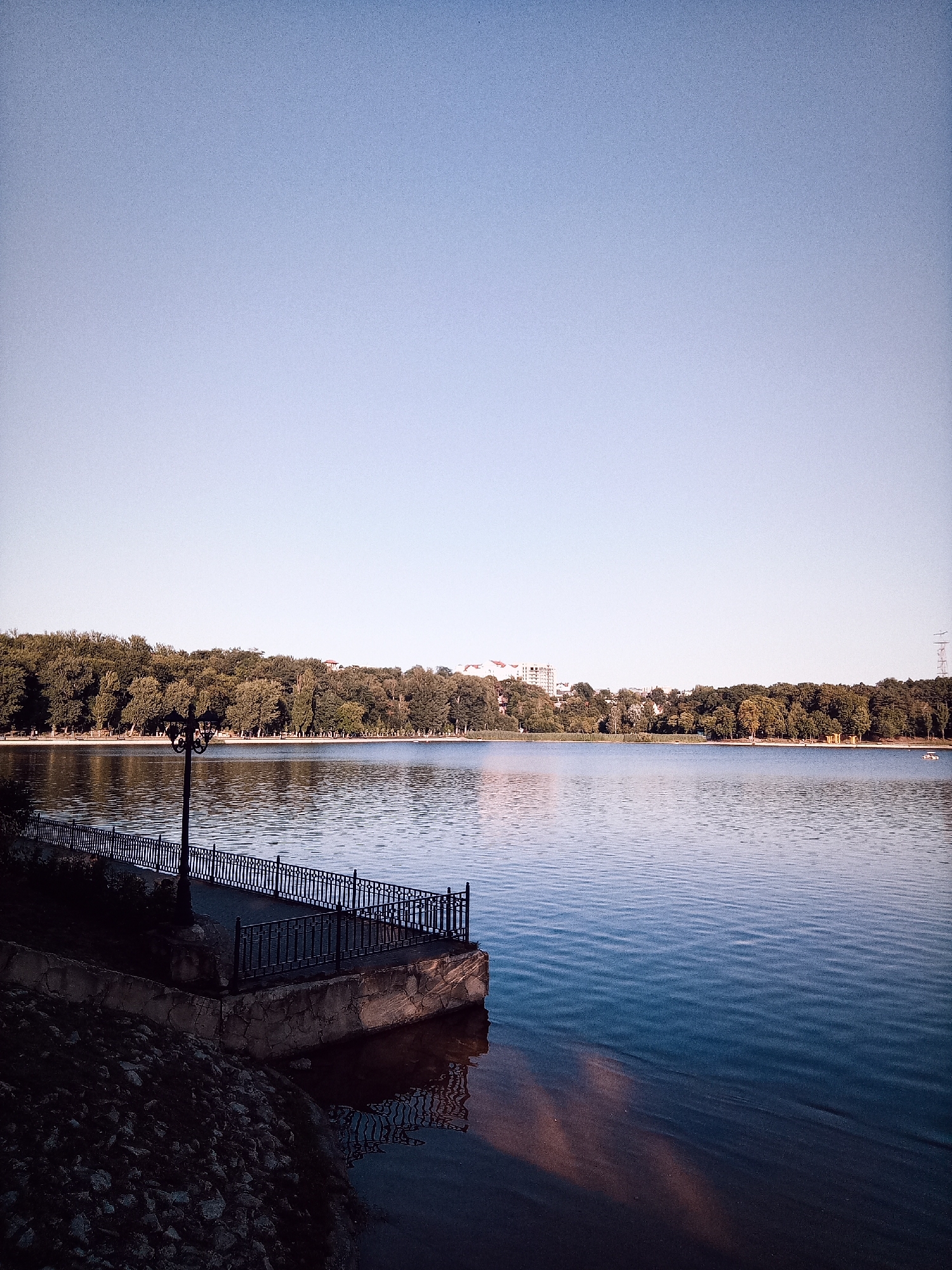The image shows a wide, calm lake with a clear sky overhead. A dense line of trees borders the far side of the lake, with a few buildings visible interspersed among the foliage. In the foreground, a stone embankment with a metal railing and a lamppost overlooks the water. The surface of the lake is smooth, with gentle ripples reflecting the sky and trees. The overall atmosphere is serene and natural. This scene is consistent with views of the Valea Morilor Lake in Chișinău, Moldova, a popular recreational area with a prominent lake and surrounding parkland. There are no people or animals clearly visible in the image.