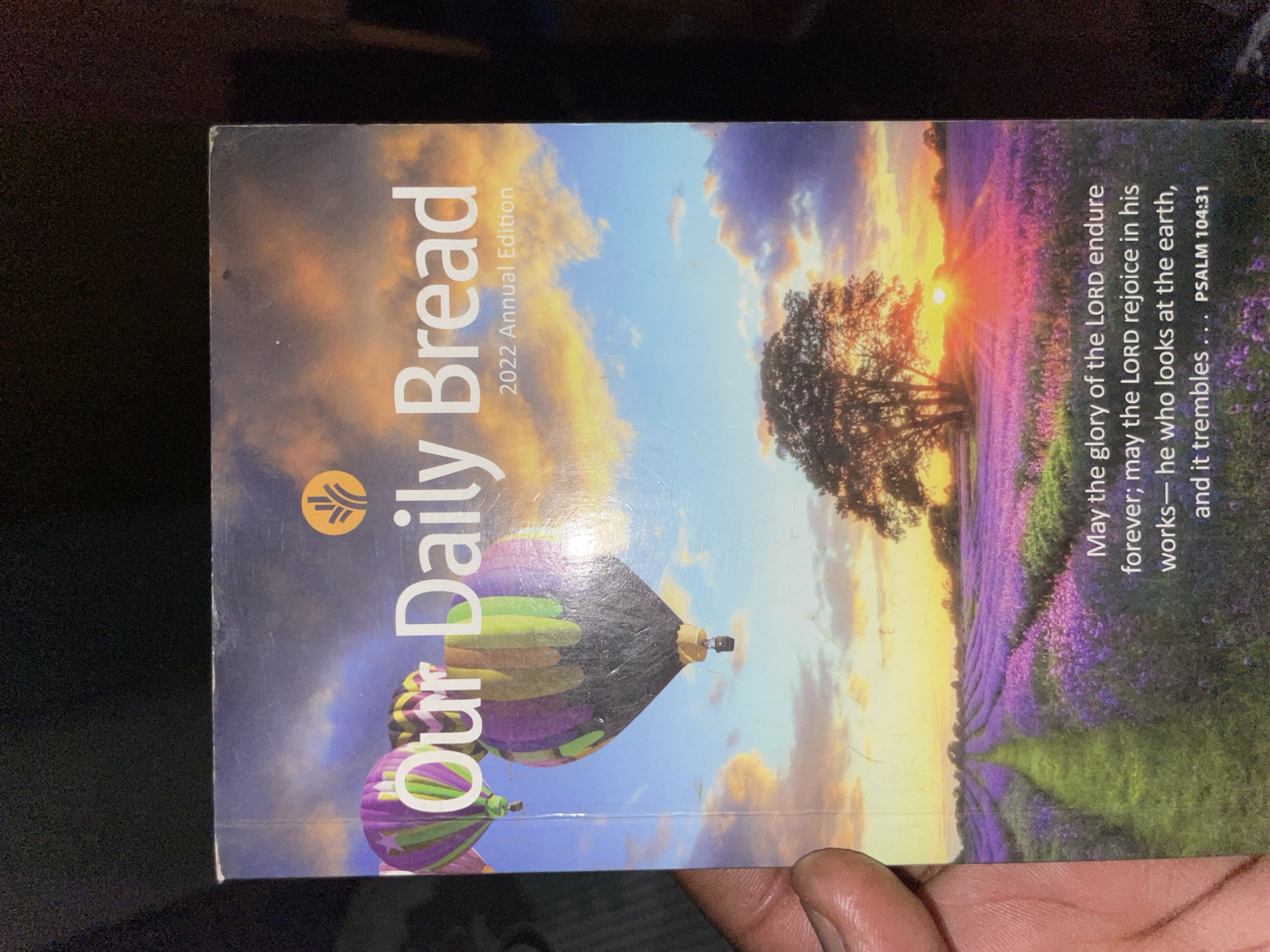 A close-up shot shows the cover of "Our Daily Bread" 2022 Annual Edition. The cover features a daytime landscape with hot air balloons, a field of lavender, a tree, and a colorful sky. The Bible verse Psalm 104:31 is printed on the right side. A human finger is visible in the lower right corner. The background is dark. The image location is unidentifiable in Kurunduma, Nigeria.