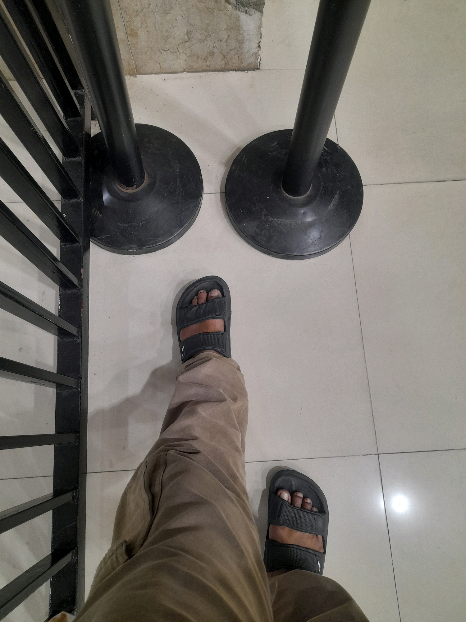 A person's feet are visible, wearing dark gray sandals and light brown pants. They are standing on a tiled floor, with two black stanchions with wide circular bases to their right. To the left of the frame is a black metal railing. The scene appears to be an indoor queuing area. The image appears to have been taken in Surabaya, Indonesia, based on the characteristic white tiled floor and the presence of stanchions often seen in public spaces like shopping malls or event venues.