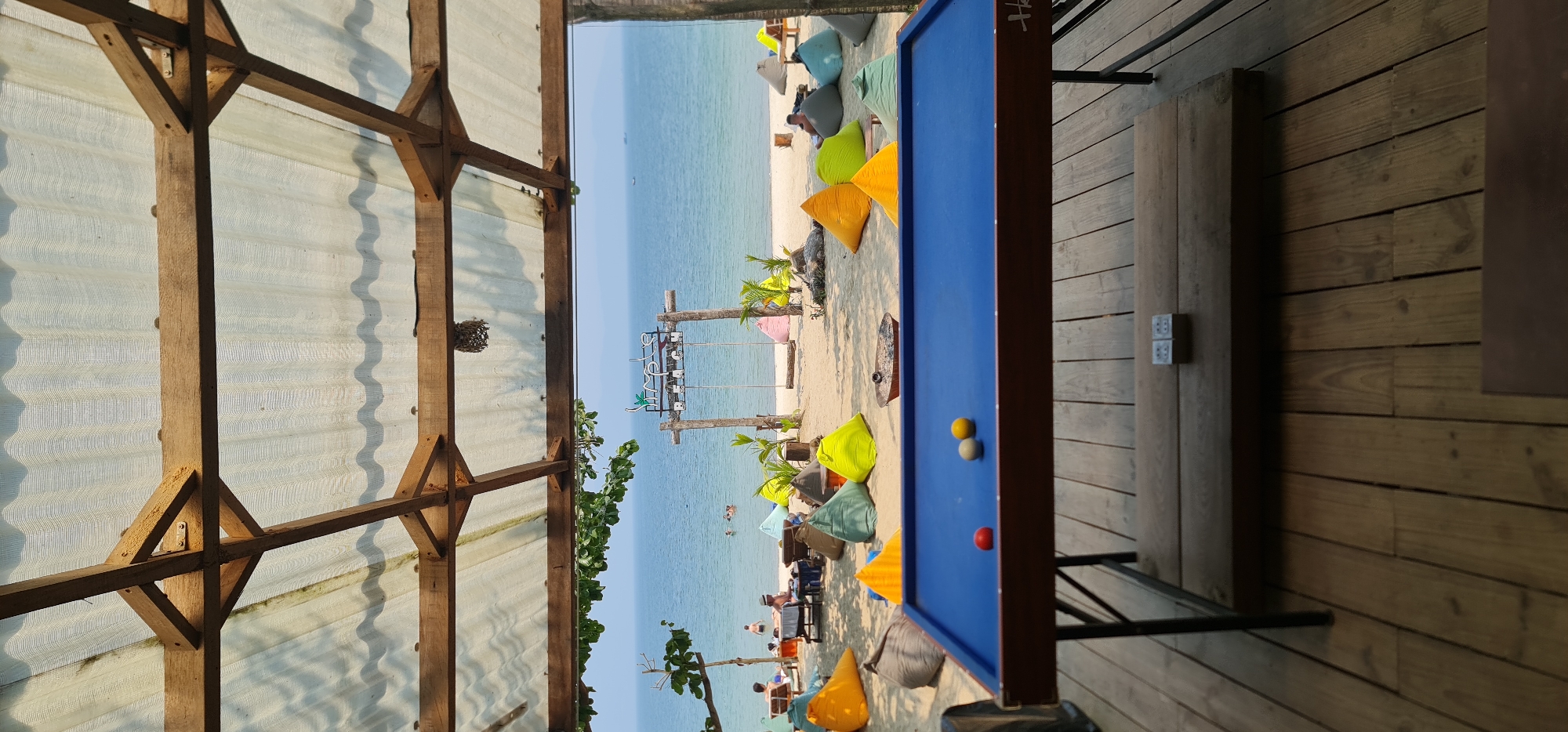 The image captures a beachside scene in Đà Nẵng, Vietnam, viewed from a partially covered structure. The structure has wooden beams and a translucent roof. Beyond this, the frame reveals a stretch of sandy beach with a clear, blue sea.
People are scattered across the beach, some sitting in colorful beanbag chairs and others wading in the water. Several trees and plants are interspersed along the beach. A decorative sign stands near the water's edge.
In the foreground, a blue billiard table stands on a wooden deck. Three billiard balls are visible on the table. The deck has an electrical outlet.