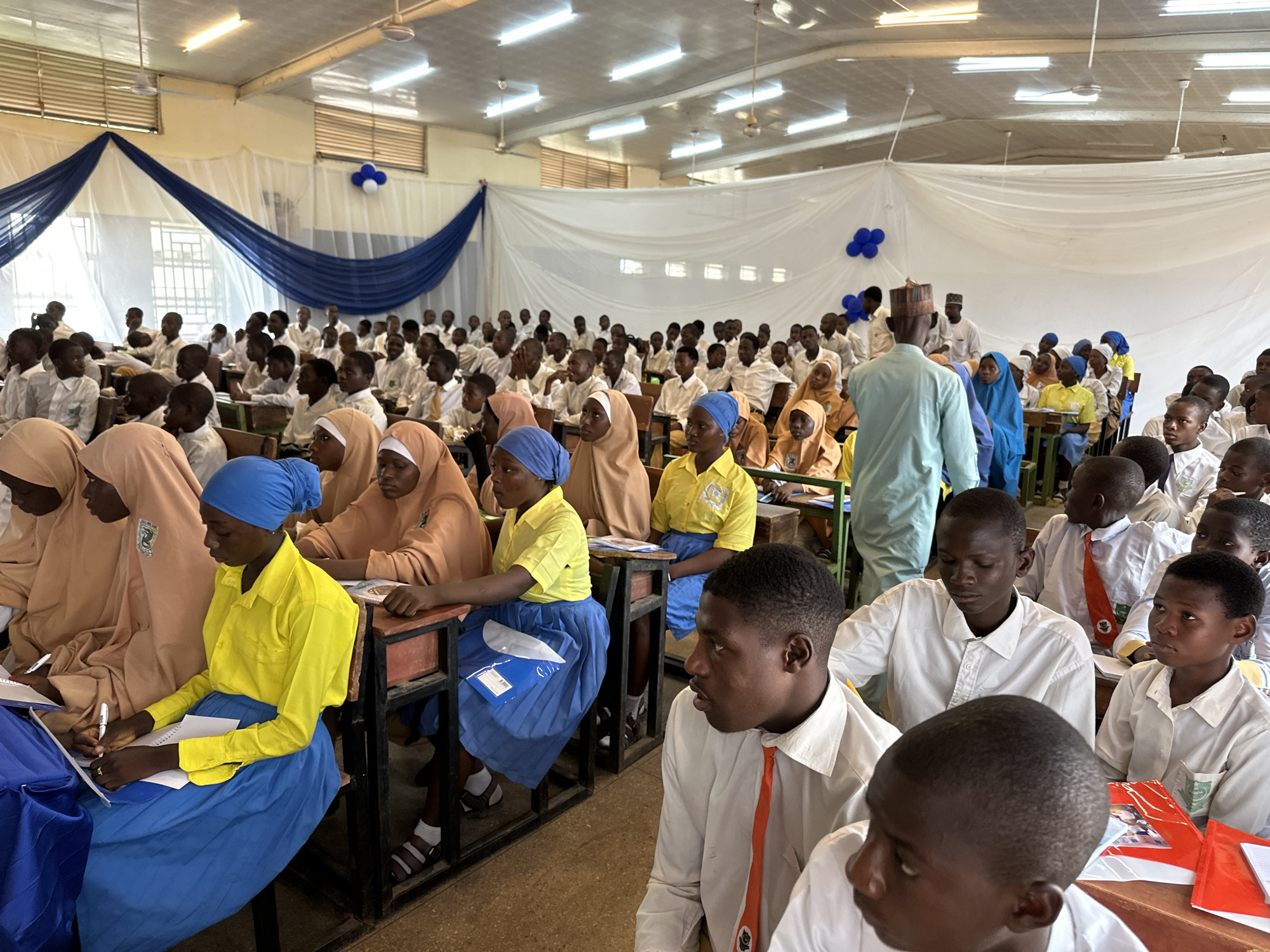 This image captures a large group of students and a few adults gathered in what appears to be a school hall or large classroom in Unguwan Dosa, Nigeria. The hall is decorated with white and royal blue fabric drapes across the back wall and blue and white balloons near the ceiling, suggesting a special event or assembly. Overhead, fluorescent lights and ceiling fans are visible.
The majority of the attendees are students, primarily teenagers, seated at wooden desks arranged in rows. Most male students wear white long-sleeved shirts, some with ties (one orange, one red). Many female students wear head coverings (hijabs or scarves) in various colors like tan, blue, and yellow. Some also wear yellow shirts and blue skirts. They appear to be attentive, looking towards the front or side, indicating they are listening to a speaker or watching a presentation. Several students have notebooks and pens, with some actively writing or looking at papers. Red and blue folders or books are also visible on desks.
An adult, dressed in a light blue traditional garment and a patterned cap, stands in an aisle with his back mostly to the camera, observing the students or preparing to address them. Other adults are visible in the background. The atmosphere seems to be one of focused engagement and learning.
While the exact time of day is not clear, the presence of both natural light from windows and artificial lighting suggests it could be daytime. A green logo, possibly a school emblem, is visible on the pocket of one student's white shirt. On a blue folder, partially visible text might read "CLIN" or "CUIN."