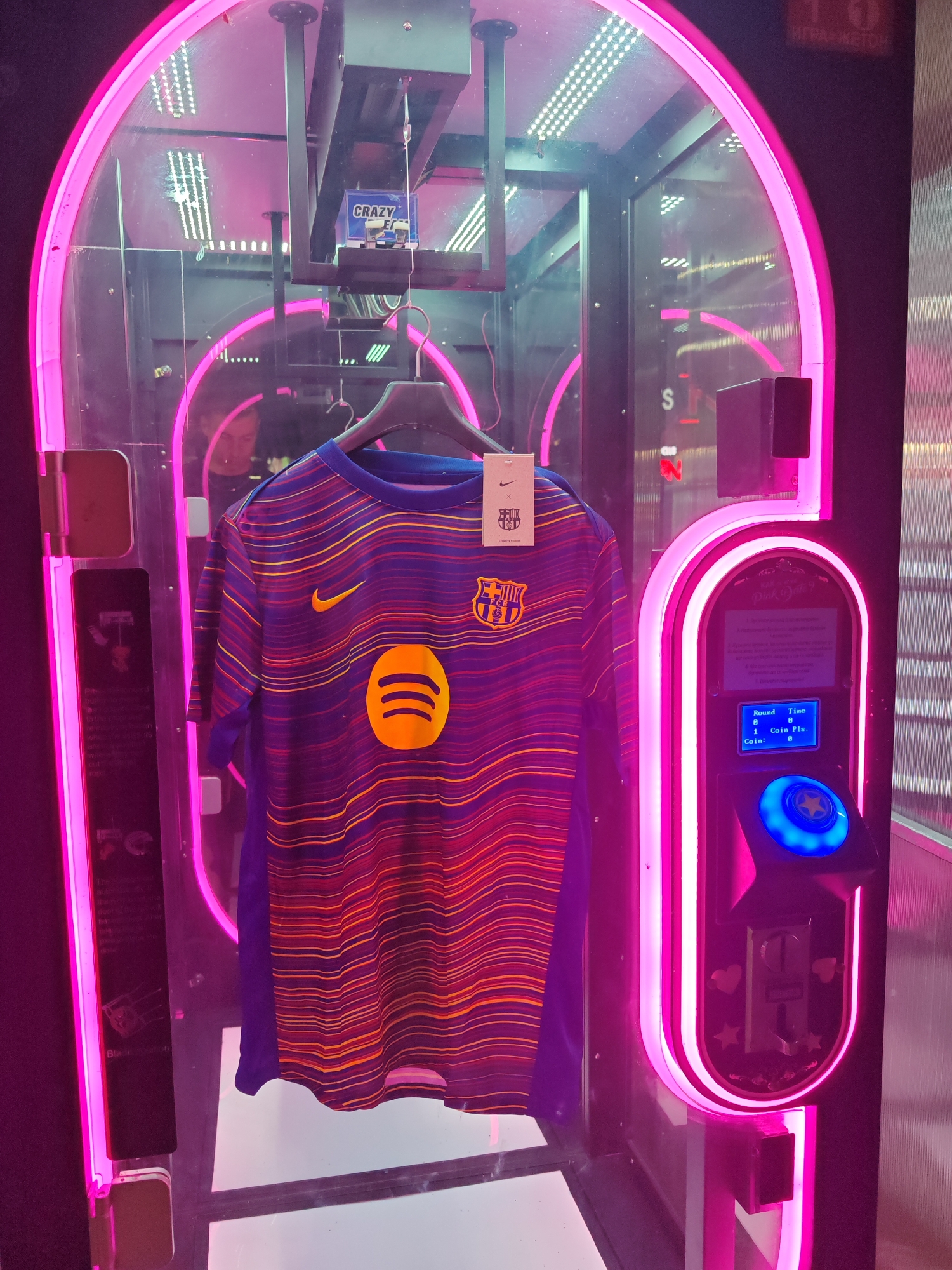 Here is a detailed description of the image:
**Scene:** A close-up of a claw machine, likely located in an arcade or entertainment venue in Nesebar, Bulgaria. The machine has a bright, neon pink frame and contains a prize of a FC Barcelona soccer jersey with the Spotify logo prominently displayed.
**People:** No people are visible directly, but a reflection of a person can be seen in the glass of the claw machine.
**Subjects/Objects:**
* **Claw Machine:** Illuminated with neon pink lights, control panel visible on the right side, containing a screen with instructions.
* **Barcelona Jersey:** A FC Barcelona soccer jersey is hung inside the machine on a hanger, likely the grand prize. It has the Barcelona emblem, a Nike swoosh, and the Spotify logo.
* **"CRAZY" Box:** A box with the word "CRAZY" is visible in the upper portion of the machine, possibly part of the claw mechanism or decorative elements.
* **Control Panel:** Has a screen displaying "Round Time 0 0 1 Coin Pls."
* **Signage:** Some text on the side of the machine on a sign board.
**Environment:** The environment appears to be an indoor gaming area or arcade, based on the lighting and machine-style setup. There is reflective paneling visible on the right side, suggesting other machines or arcade games are nearby.
