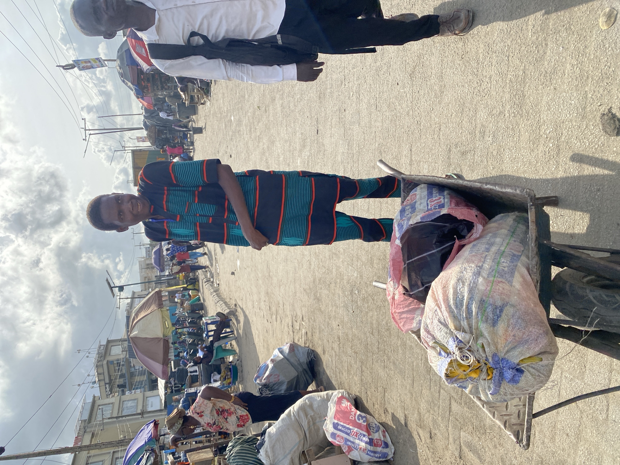 The image depicts a street scene in Lagos, Nigeria. A young man in a striped shirt and pants stands next to a wheelbarrow filled with sacks. An older man in a white shirt stands slightly behind him. The background shows a crowded marketplace with stalls and people under umbrellas. Buildings and utility poles are visible in the distance under a partly cloudy sky. The ground is paved, suggesting a street or a walkway. The image is taken at ground level with a slight rotation.