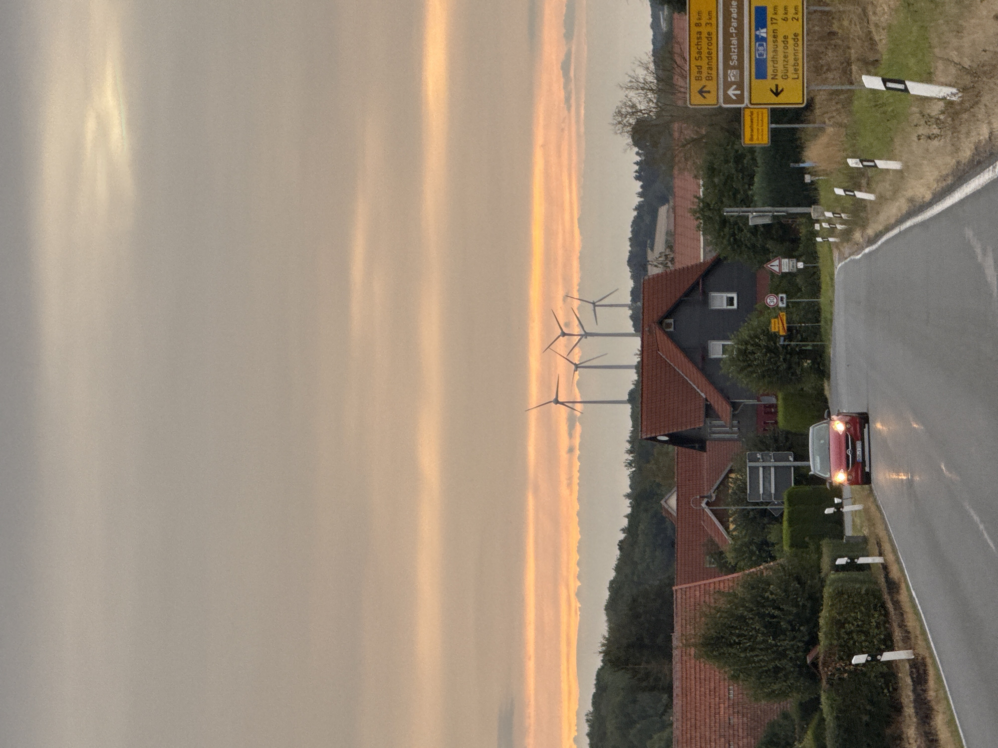 This image depicts a road in Hohenstein, Germany, with a red car driving towards the viewer. The car's headlights are illuminated, and the road appears wet, suggesting recent rain. The scene is framed by lush green hedges lining the road and several houses with red tiled roofs. In the background, wind turbines are visible against a sky with a warm, orange hue, indicative of sunset or sunrise. Several yellow road signs are present, providing directional information to various locations, including Bad Sachsa, Branderode, Nordhausen, Günzerode, and Liebenrode. The overall environment suggests a rural or semi-rural setting within Hohenstein.