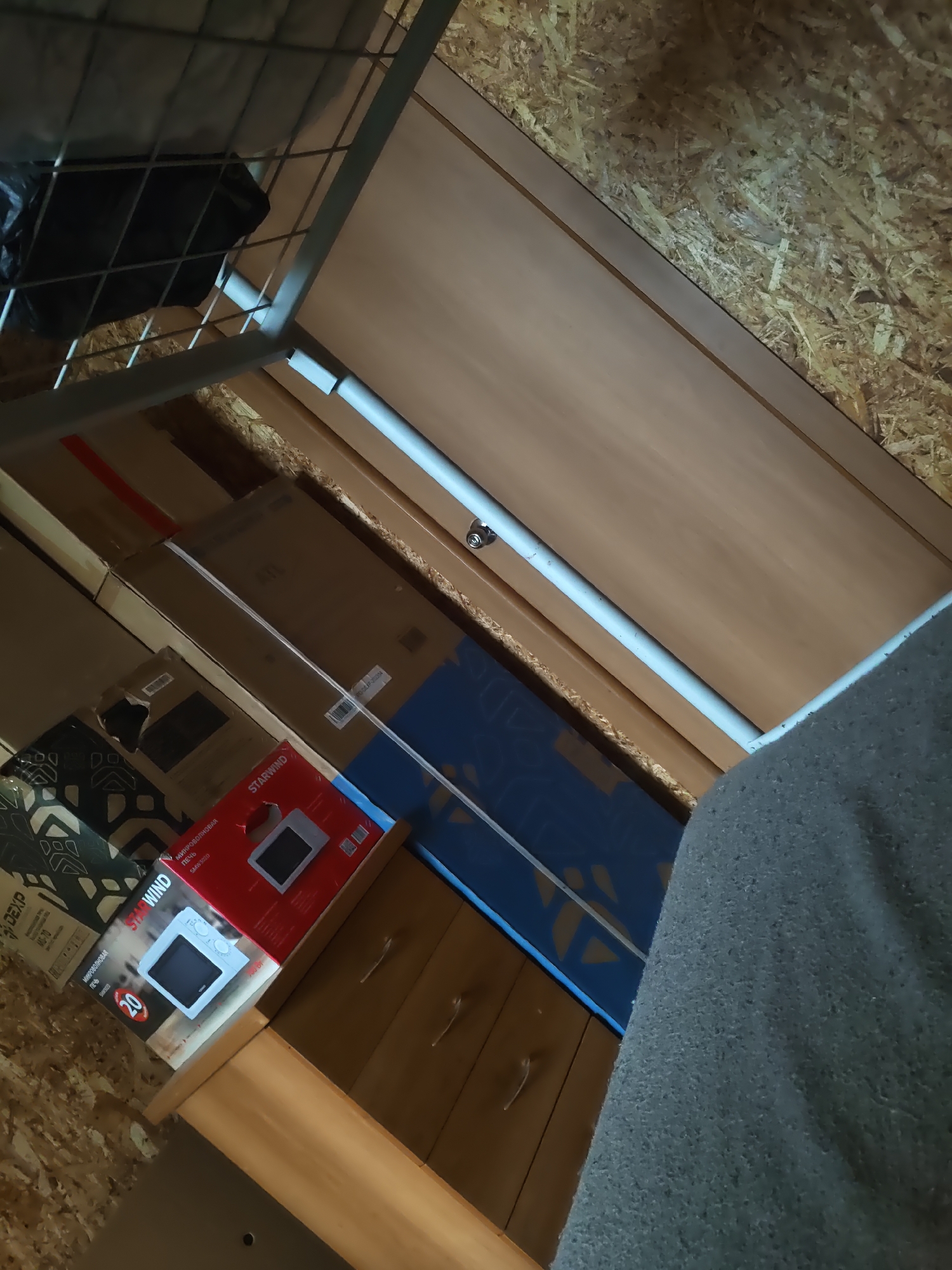 The image shows a cluttered storage area with several boxes and a metal shelf. In the foreground, a red and white box with the brand "STARWIND" and text in Russian for "microwave oven" is visible. A white microwave is depicted on the box. To the left of the microwave box, another box with a black and white geometric pattern is partially visible, along with a box labeled "DEXP". Behind these boxes, a large blue box with a yellow pattern is stacked. A metal shelf with a gray mattress underneath is positioned above these items. The walls are constructed of OSB (Oriented Strand Board) and a light-colored wooden panel. The scene appears to be indoors. The location is identified as Platoshino, Russia.