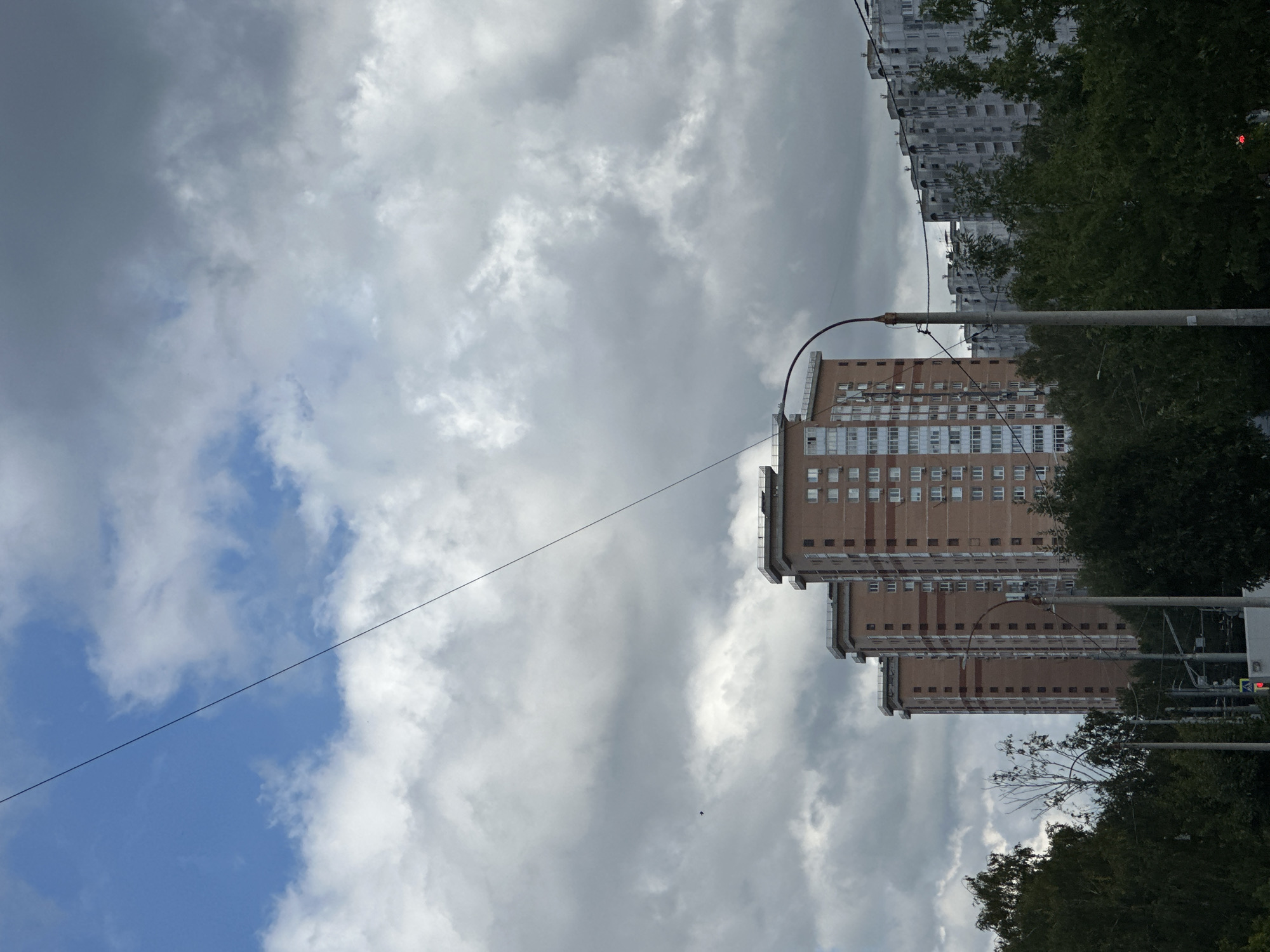 The image shows a view of apartment buildings in Podolsk, Russia, against a cloudy sky. In the foreground, a power line stretches diagonally across the frame. Behind it, several tall apartment buildings are visible. The buildings on the left are made of reddish-brown brick with many windows, while those on the right are lighter in color and appear to be made of concrete. Lush green trees line the lower portion of the image, partially obscuring the lower levels of the buildings. A street lamp with a curved arm stands in the middle ground, and traffic lights are visible in the lower left corner. The sky is filled with a mixture of blue patches and heavy, grey clouds.