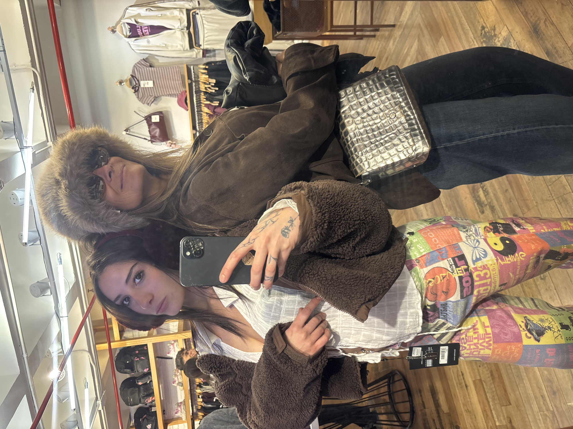 A young woman with long brown hair holds a smartphone in her right hand, angled to capture a selfie-style photograph reflected in a mirror. She is wearing a light-colored blouse, a brown sherpa jacket, and multi-colored patterned pants. Her left hand is visible near the bottom of the frame. Her skin is fair with tattoos on her left hand and wrist.
Behind and above her, an older woman wears a fur hat and sunglasses. She is wearing a brown suede jacket, a silver bag, and jeans. The mirror reflects the interior of a clothing store, featuring racks of clothes and other accessories on display. The floor is light-colored wood. There is a radiator along the left side of the mirror. The image appears to be taken indoors. Without additional visual information or context, pinpointing the exact location within Buenos Aires is not possible.