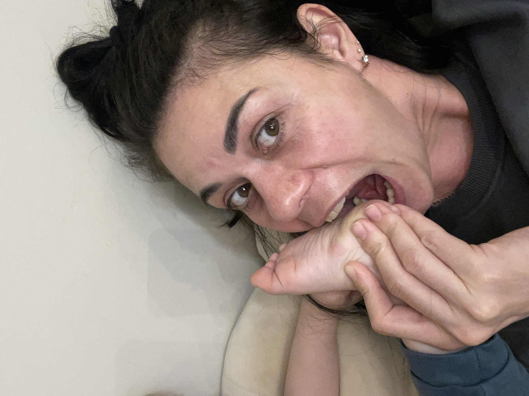 The image displays an adult female in a close-up, high-angle shot within an indoor environment in Unknown, Unknown. The subject, with dark hair pulled back, looks directly at the camera with wide, brown eyes and an open mouth revealing upper and lower teeth. She wears a dark-colored, long-sleeved garment. Her right hand grasps a bare human foot, positioning its sole near or within her open mouth. A segment of a leg, presumably belonging to the foot, is partially visible in the upper left portion of the frame. The background consists of a plain, light beige surface, likely a wall or upholstered furniture. The scene depicts a direct physical interaction between the female subject's mouth and hand, and another person's foot.